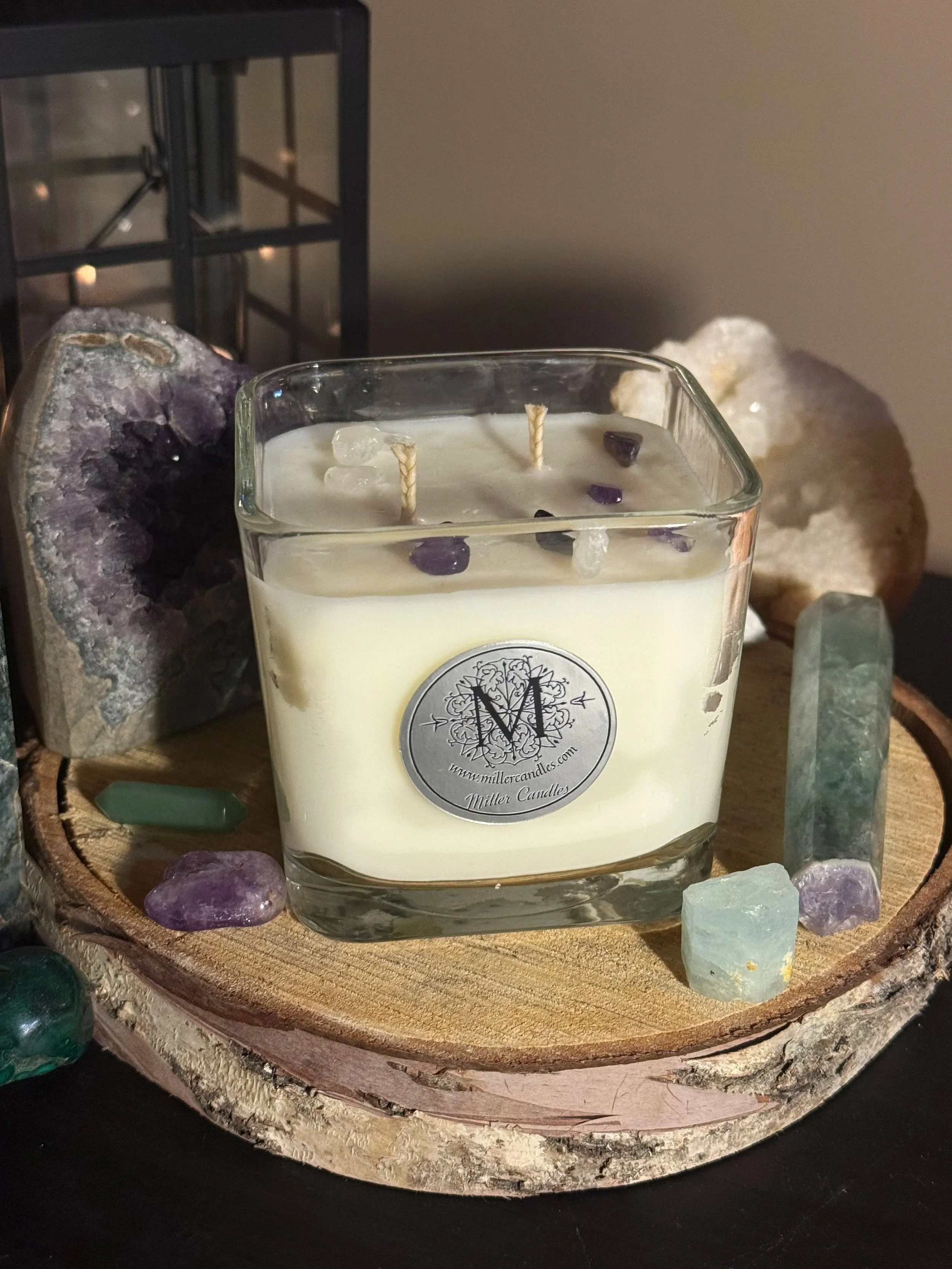 A white candle in a square glass container with three wicks, sitting on a round wooden slab, surrounded by various crystals and minerals, with a black metal lantern in the background.