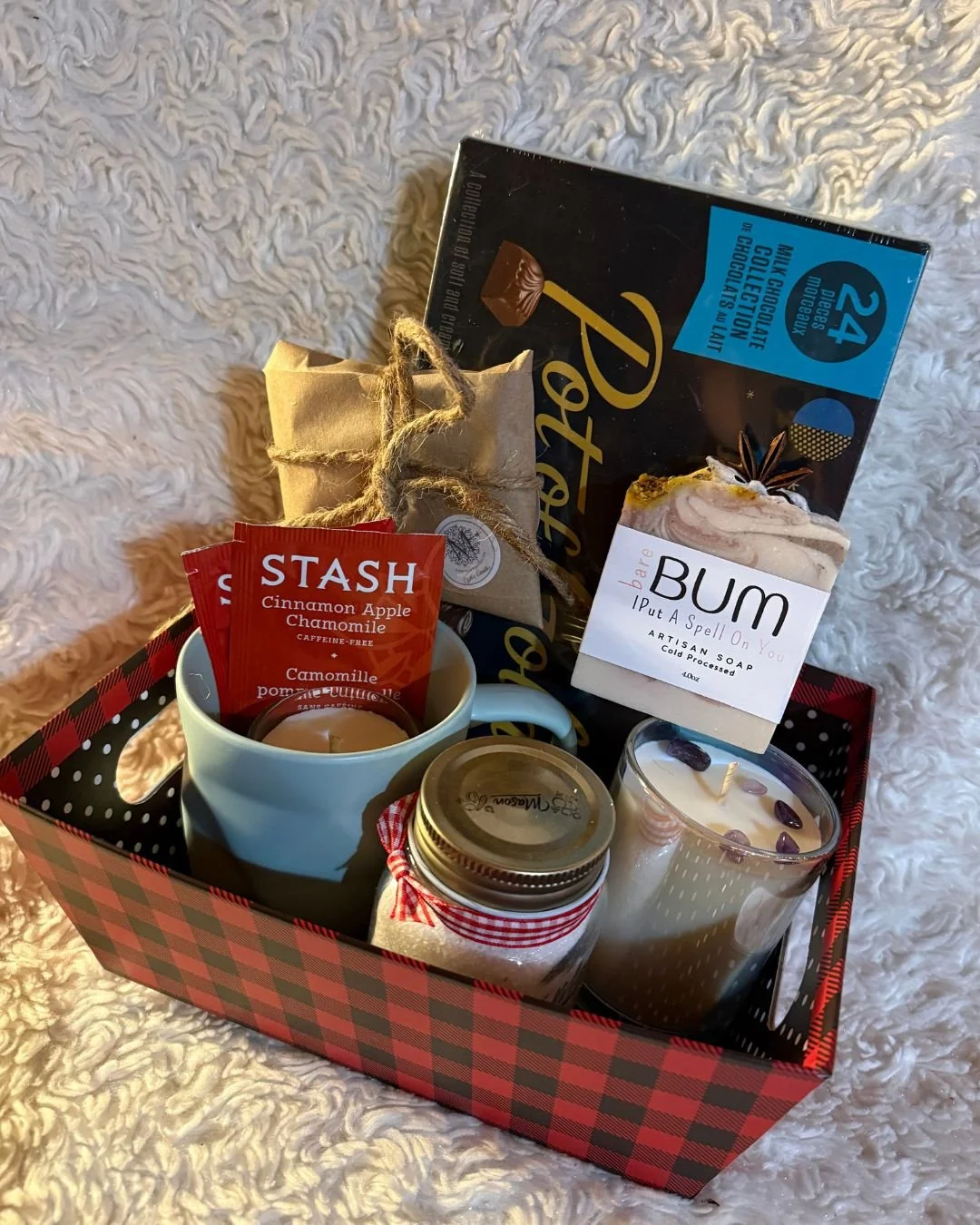A basket contains candles in glass holders, a red-orange box of cinnamon apple chamomile herbal tea, and a red, orange, and gray crocheted hat or scarf placed on a wooden and tile surface.