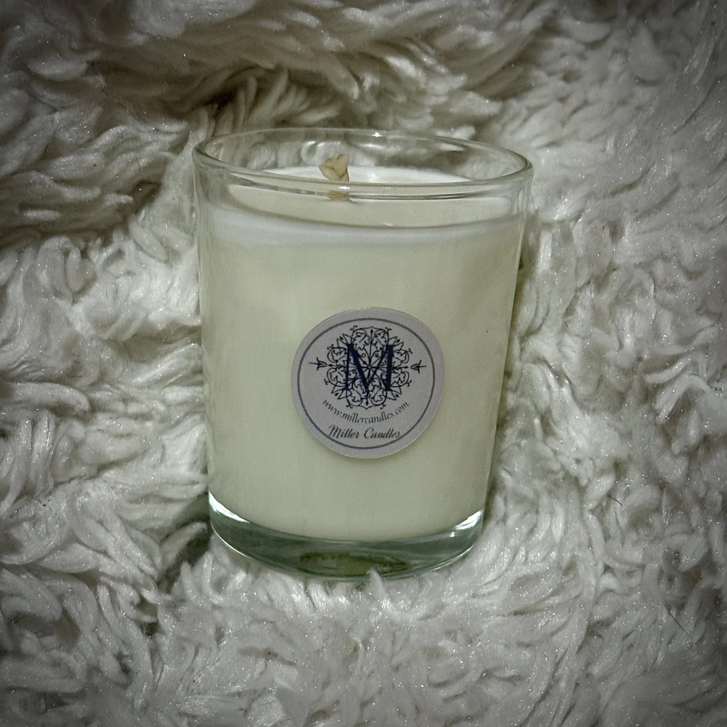 A white candle in a glass holder with a label reading 'Miller Candles' resting on a fluffy, textured white blanket.