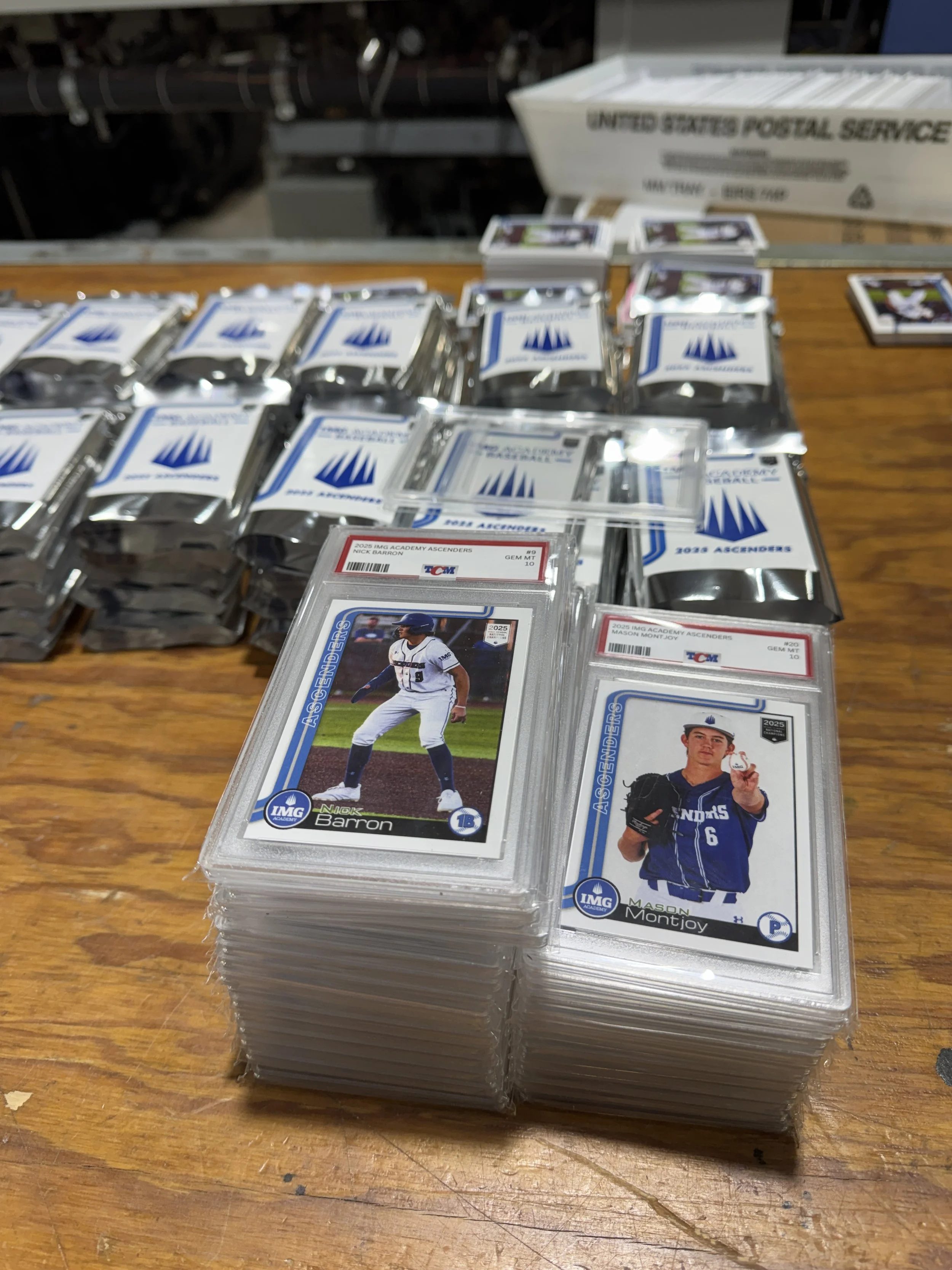 Baseball trading cards stacked on a wooden surface, with some cards featuring players Nick Barron and Mason Montjoy. Packs of cards are in the background, some labeled "2018 AACADMY ASCENDERS."
