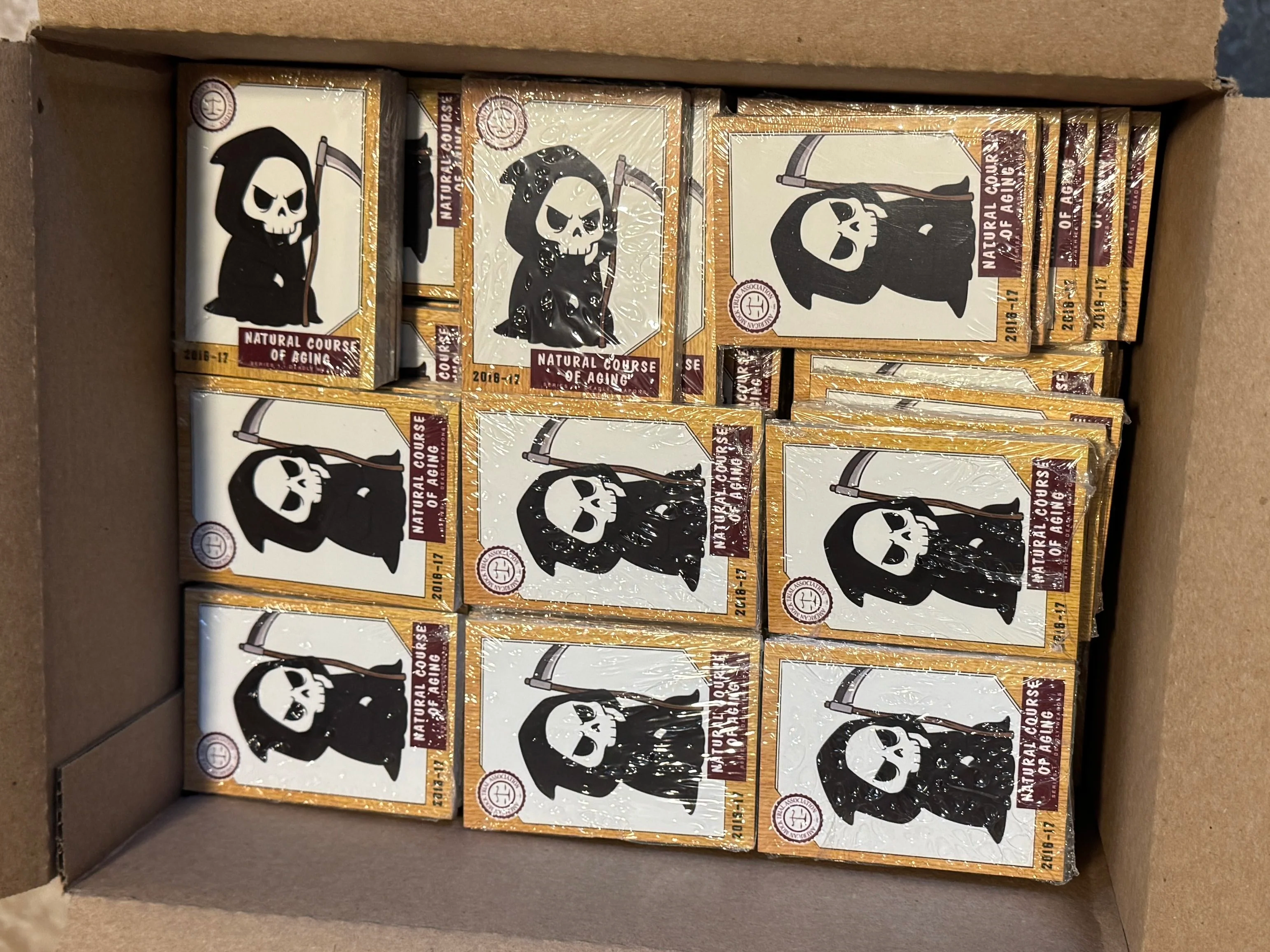A box filled with multiple packs of matches featuring skull and Grim Reaper cartoon illustrations and the label 'Natural Course of Aging'.