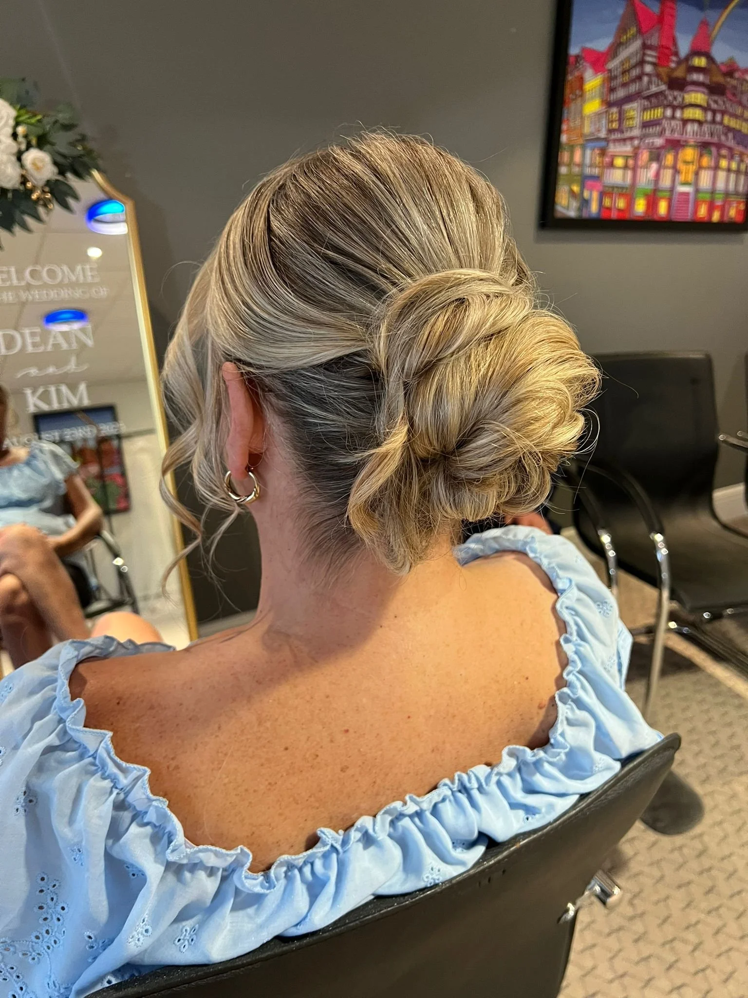 Back view of a woman with blonde hair styled in an elegant updo, sitting in a salon chair wearing a light blue, ruffled blouse.