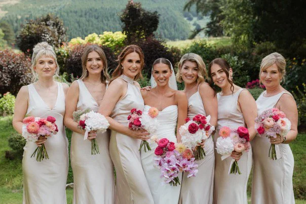 Group of seven women in white dresses at a garden wedding, holding bouquets of pink and white flowers.