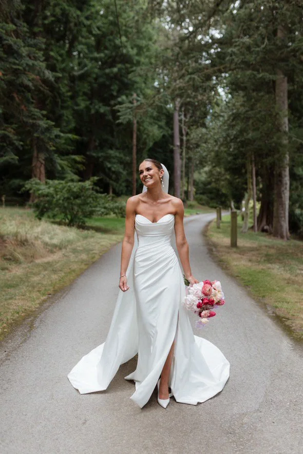 Bride in a white wedding gown holding a bouquet of pink and white flowers, smiling on a rural road surrounded by trees.