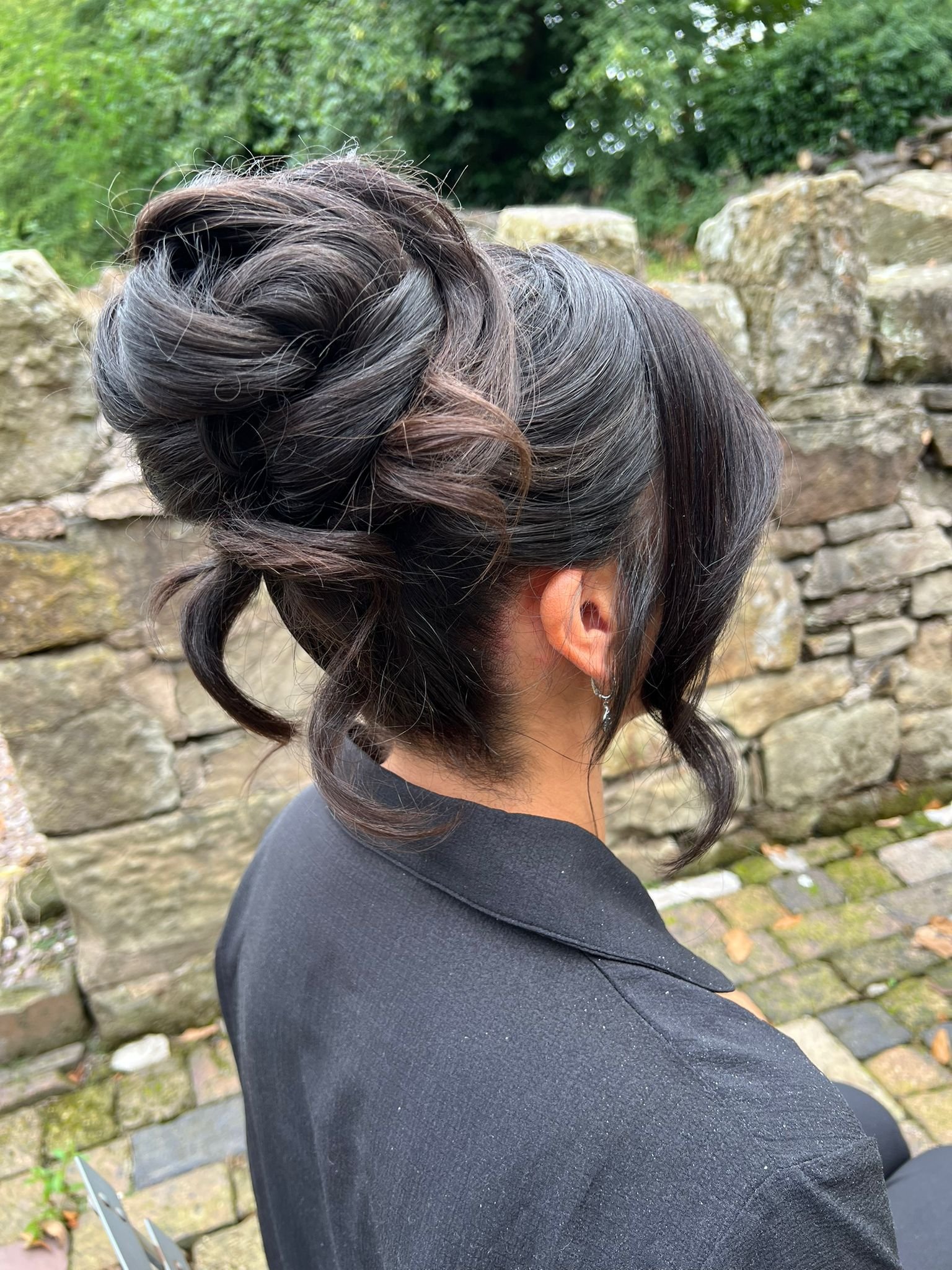 A woman with dark, styled hair in an updo, wearing a black blazer, standing outdoors near a stone wall and greenery.