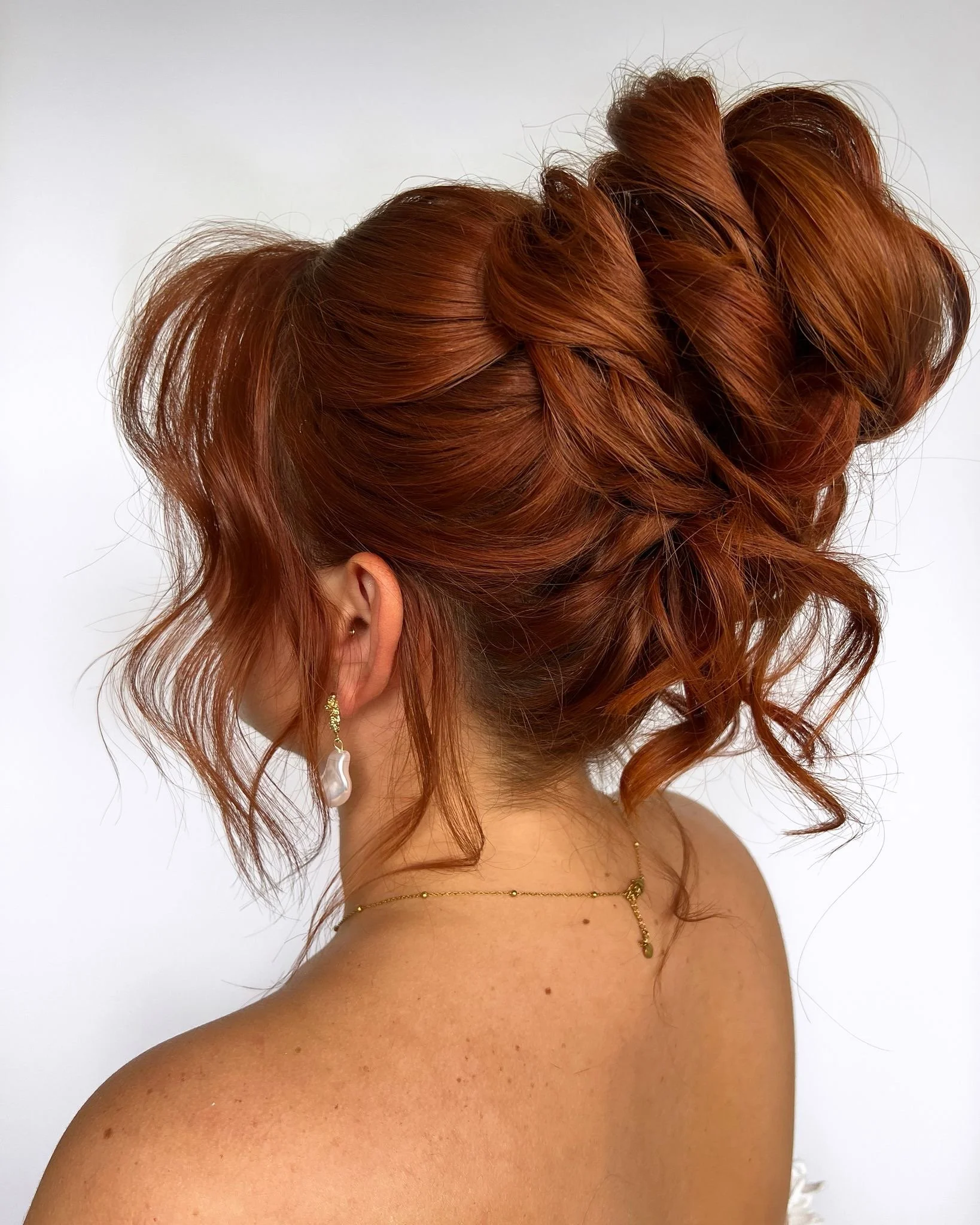 Side view of a woman with elaborate red hair styled in an updo with loose, curly strands, wearing gold jewelry and a white pearl earring, against a plain white background.