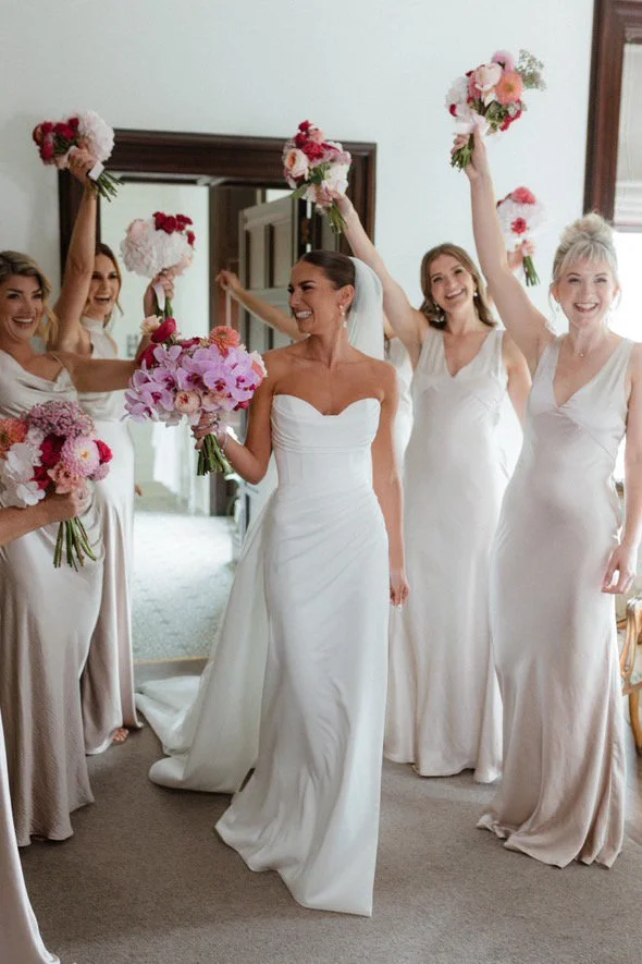 Bride and bridesmaids in white dresses celebrating with bouquets in a room with a mirror