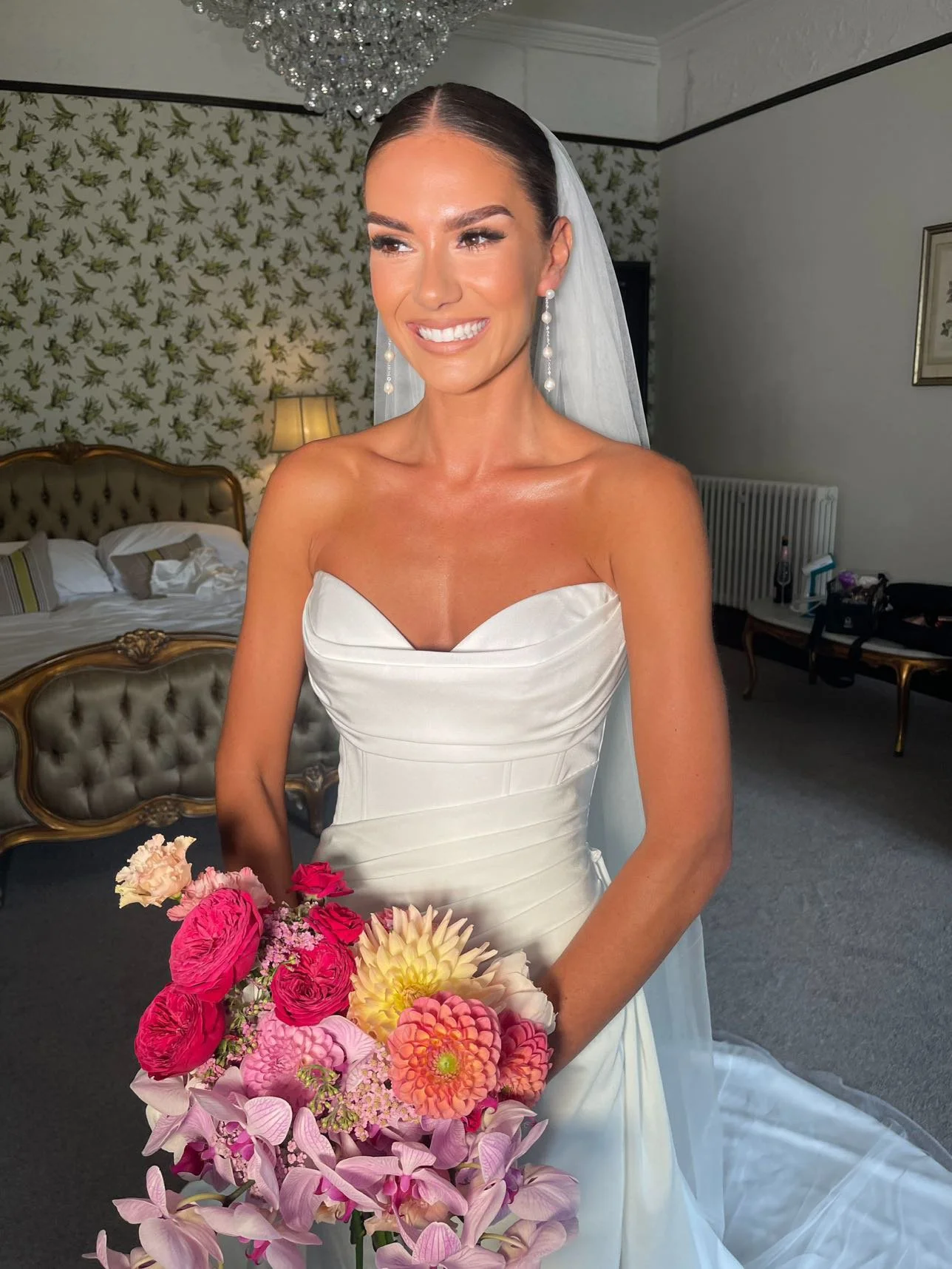 A smiling bride in a white wedding dress with a veil, holding a colorful bouquet of flowers, inside a decorated room with wallpaper, a chandelier, and furniture.