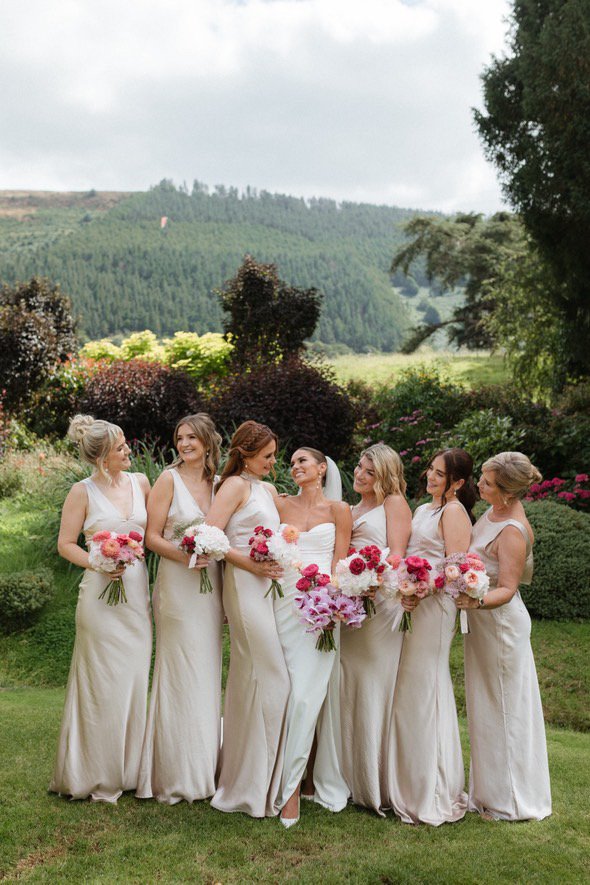 A bride and six bridesmaids standing outdoors on a grassy area with trees and hills in the background, all holding bouquets of flowers.