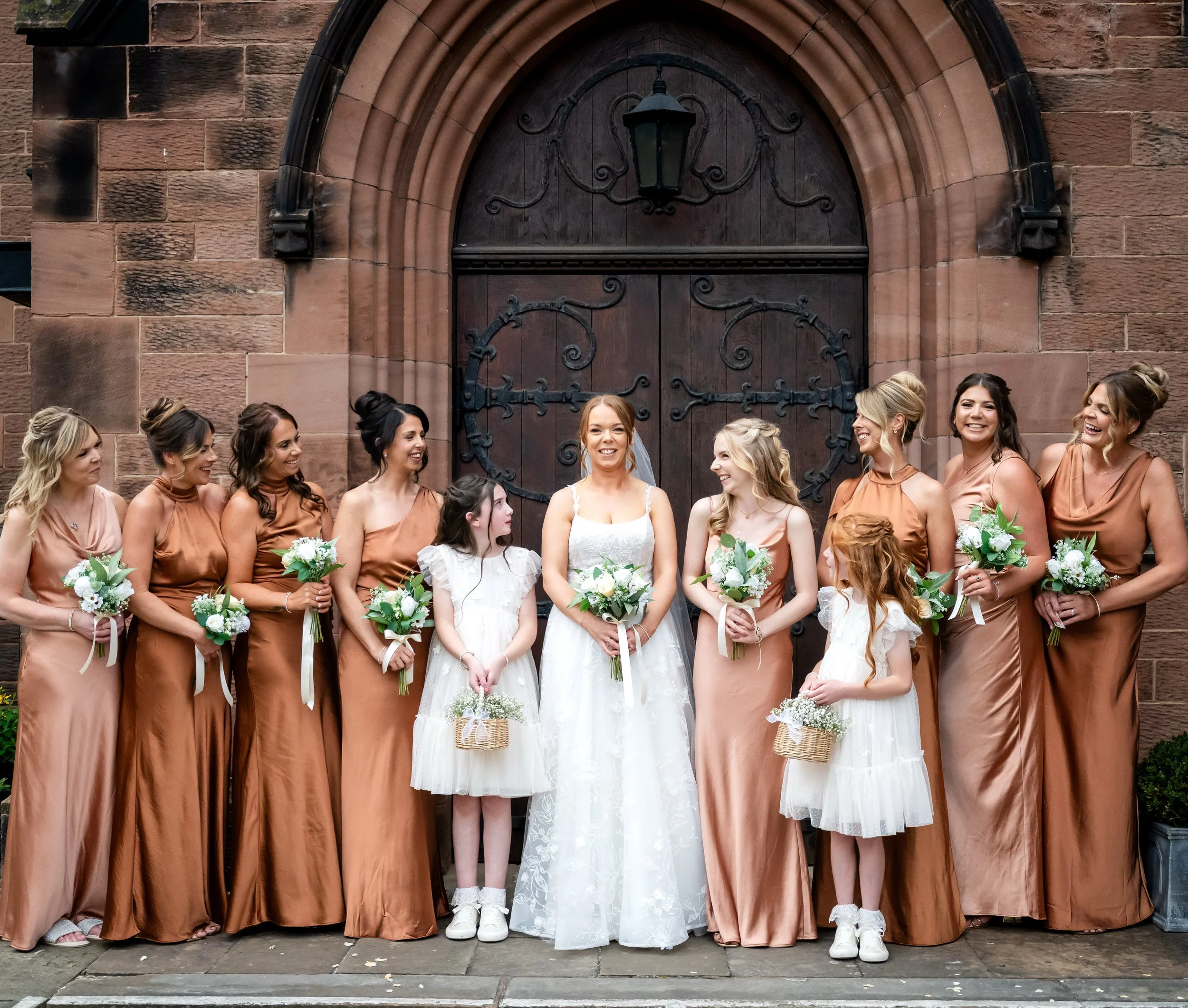 A bride and her bridesmaids standing in front of a wooden door, holding bouquets, with two young girls in white dresses holding baskets, all smiling and dressed in formal attire.
