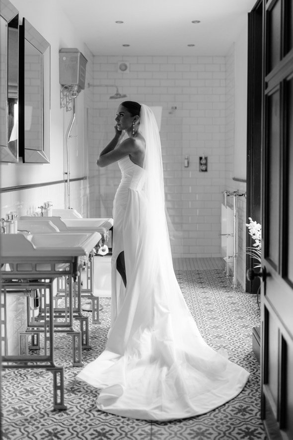 A bride in a wedding gown standing in a bathroom, adjusting her earring and looking at her reflection in a mirror.