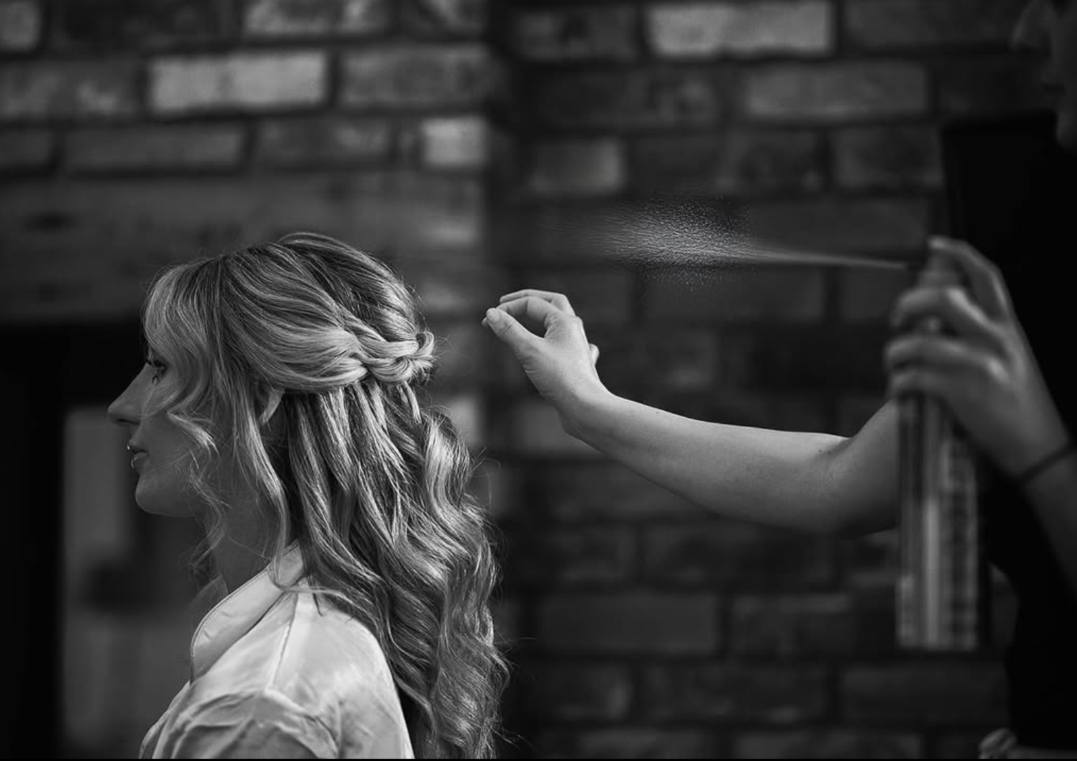 A woman with long, wavy hair styled in loose curls and a braid, is sitting in a salon while a hairstylist sprays her hair with a spray bottle. The background has a brick wall.