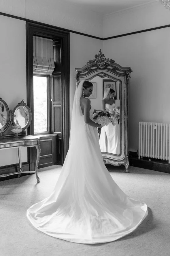 Bride in a wedding dress holding a bouquet standing in front of a mirror in a vintage room with a large window, ornate furniture, and a full-length mirror.