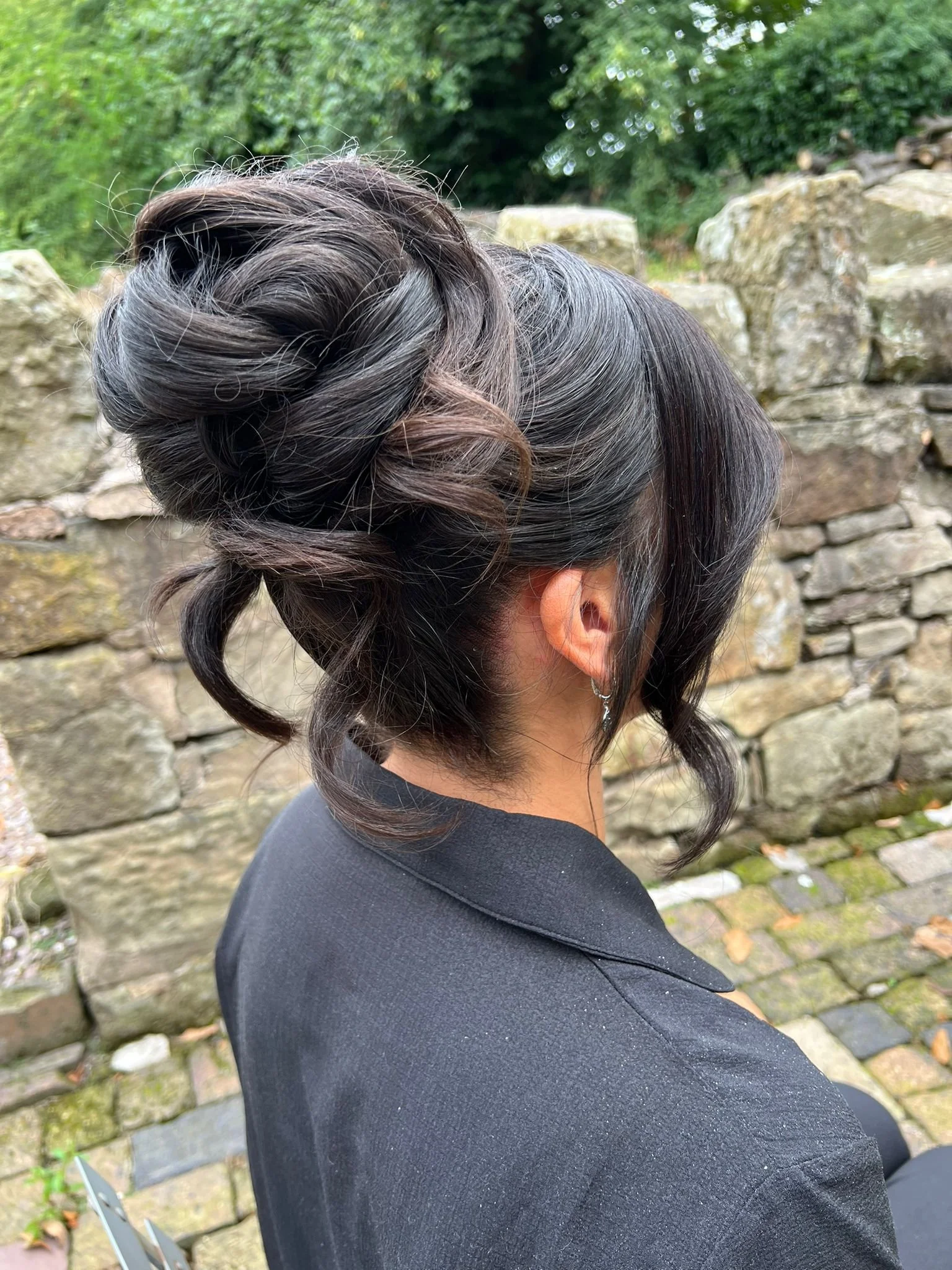 Side view of a woman with dark, wavy, styled hair standing outdoors in front of a stone wall and green foliage, wearing a black jacket and hoop earrings.
