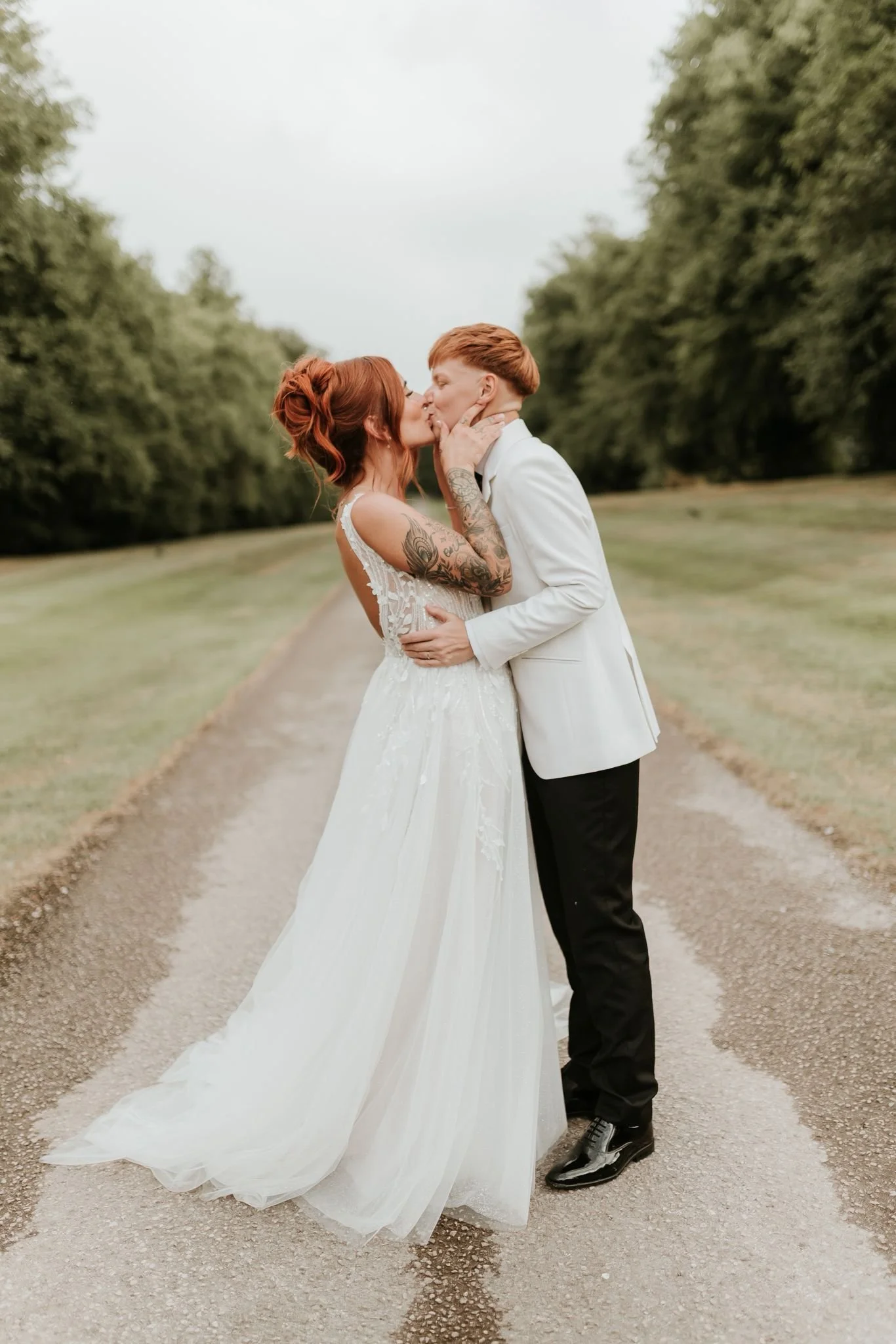 A couple in wedding attire sharing a kiss on a gravel road surrounded by trees.