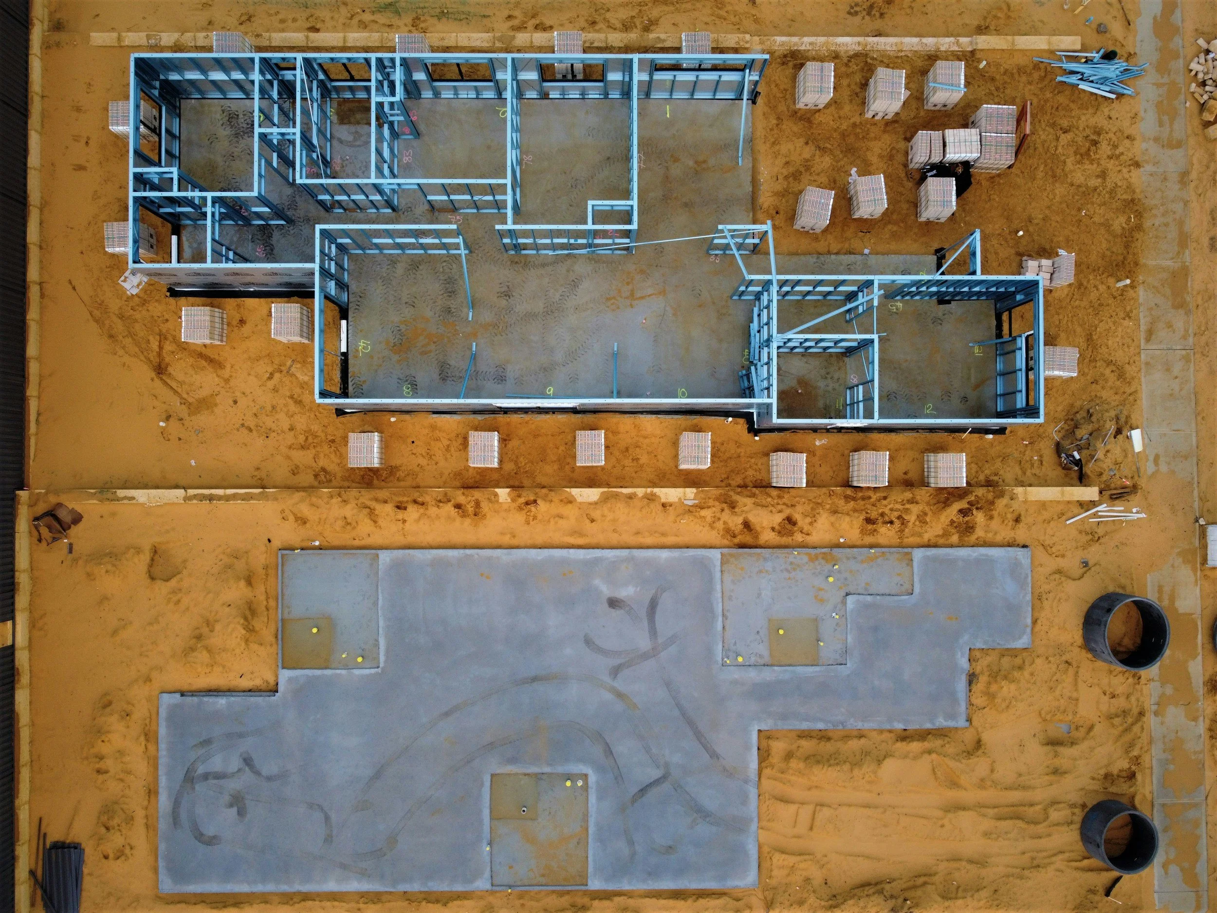 Aerial view of a building under construction with blue steel framing, surrounded by sandy ground, with a concrete pad, and construction materials and tools nearby.