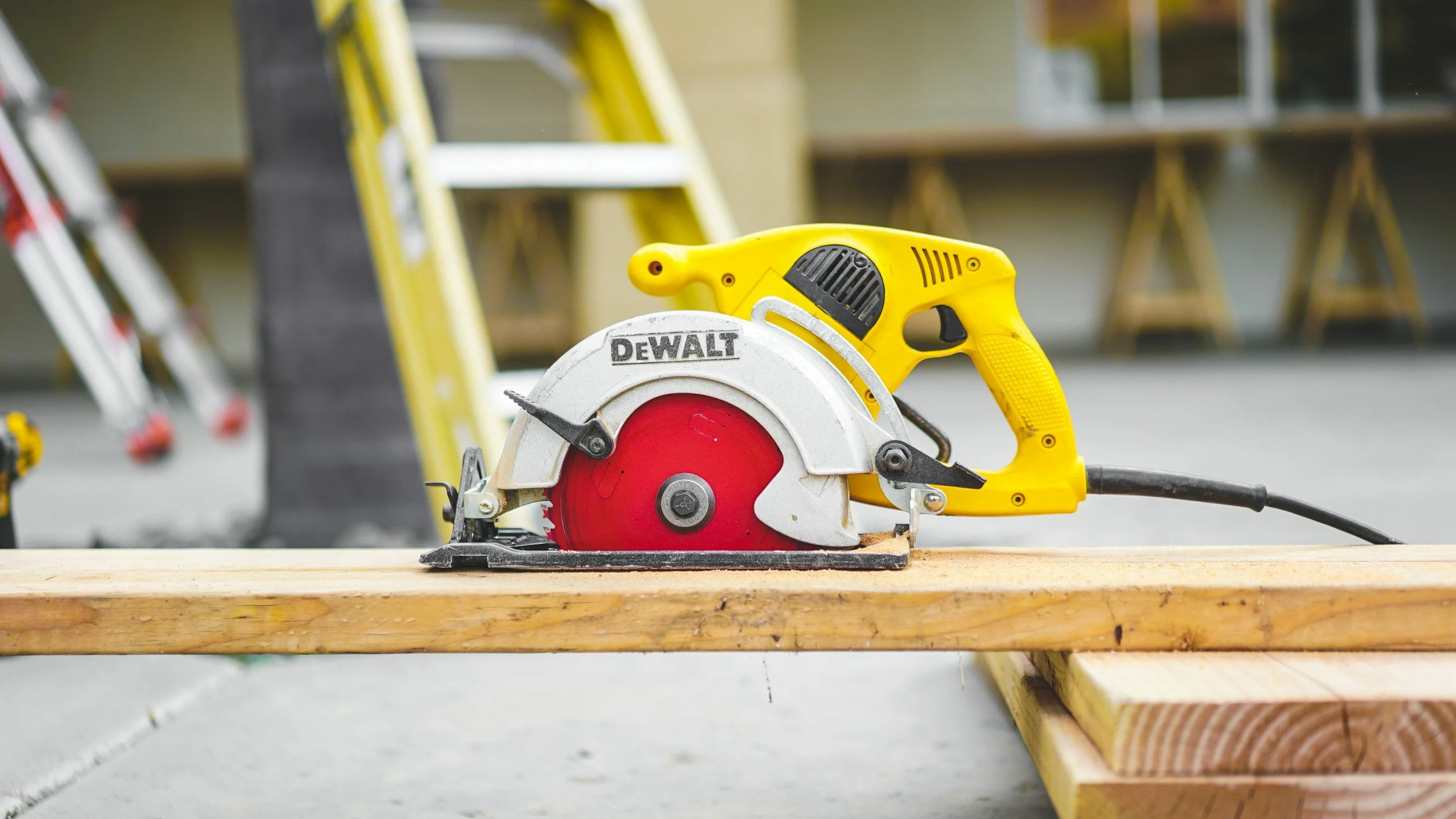 A yellow DeWalt circular saw placed on a piece of wood at a construction site with ladders and tools in the background.