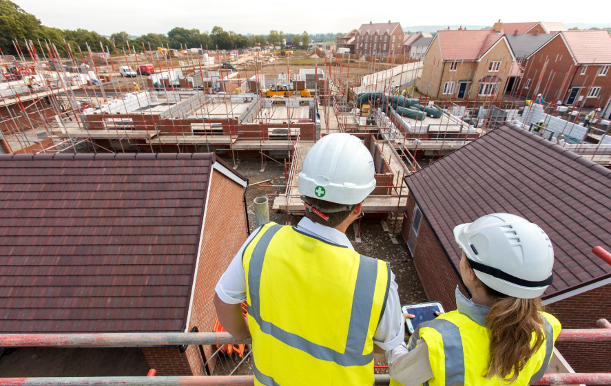 Two construction workers wearing safety helmets and vests overlooking a construction site with multiple buildings and scaffolding.