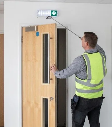 Person in a high-visibility vest cleaning an emergency exit sign above a door with a long duster in an indoor hallway.