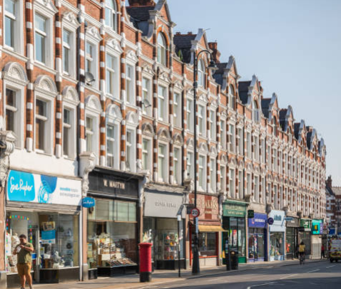 A street with historic buildings featuring shops on the ground level and residential apartments above, with pedestrians walking along the sidewalk.