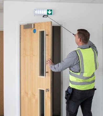 A man in a yellow safety vest cleaning a door with a retractable duster near an emergency exit sign.