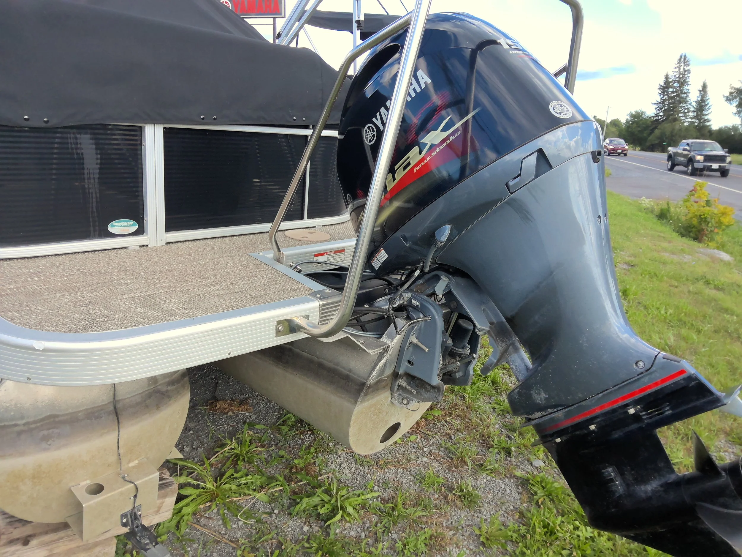 Close-up of a boat's outboard motor, specifically a Yamaha four-stroke engine, attached to the boat's stern. The boat is on a trailer, and part of the vehicle's wheel is visible. The background shows a parking lot with cars and some greenery, under a partly cloudy sky.