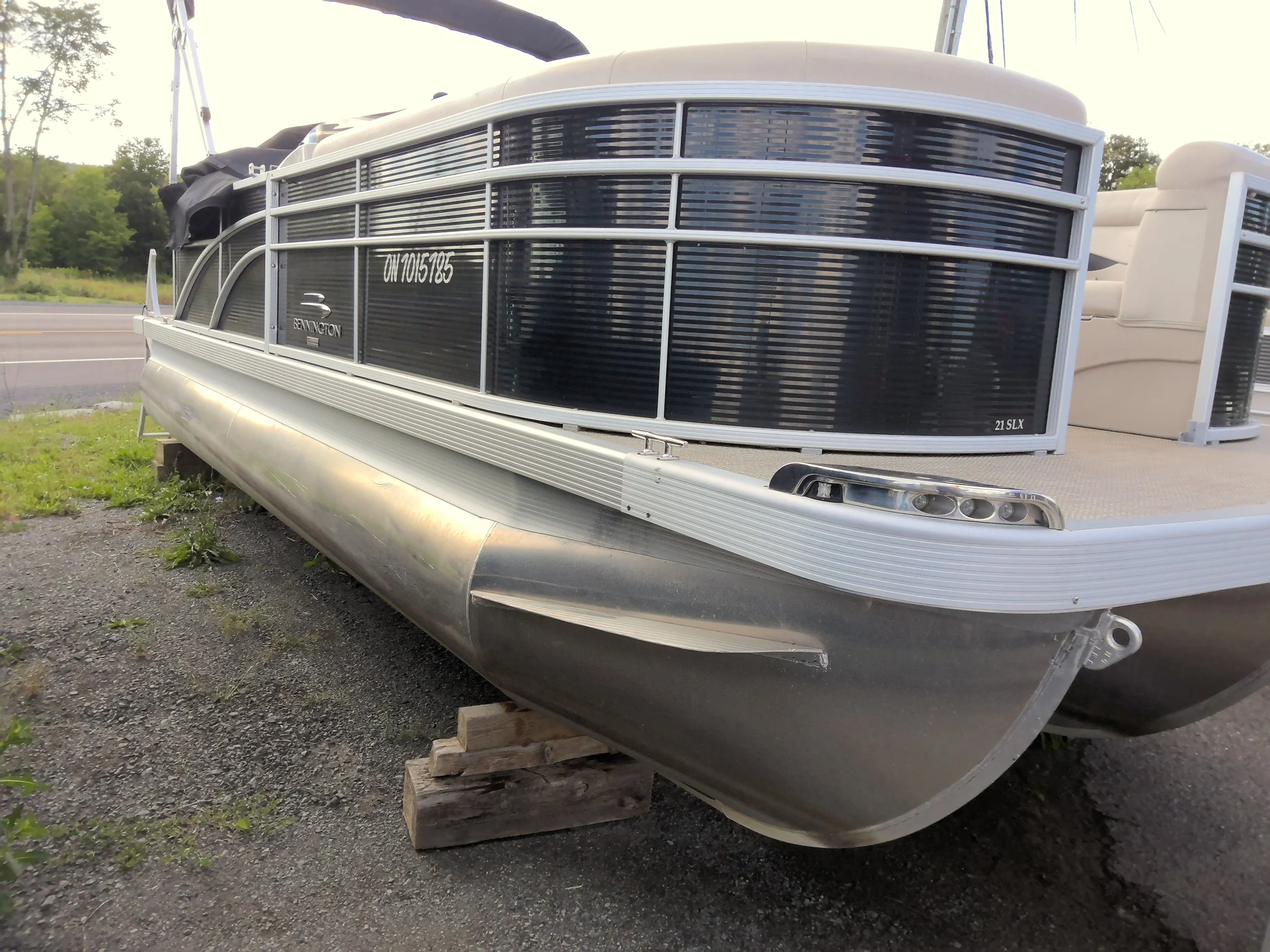 A pontoon boat resting on wooden blocks on a gravel area near a parking lot, with trees in the background.