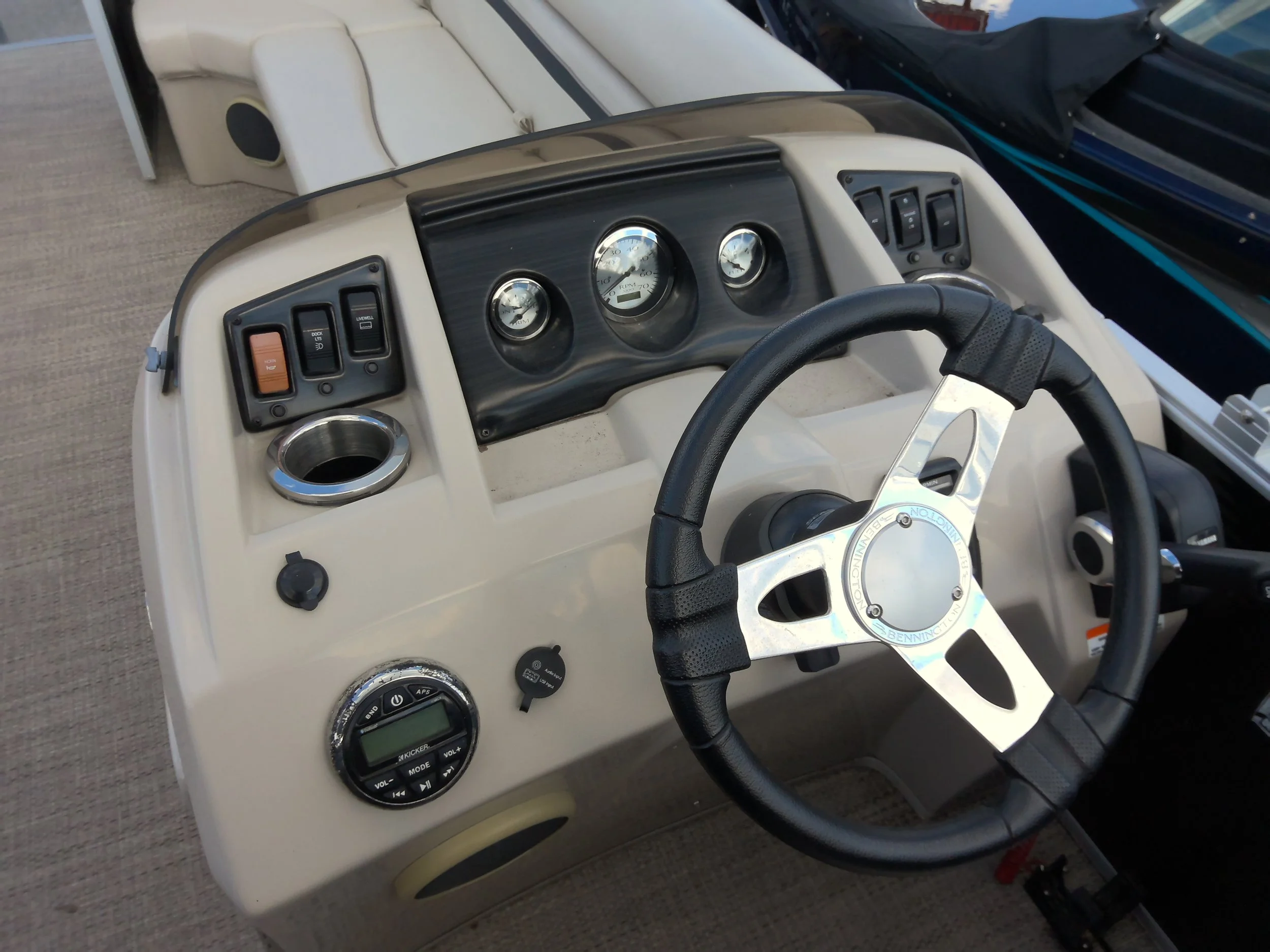 Interior of a boat with a steering wheel, three circular gauges, a cup holder, and several control switches, with part of the windshield and another vehicle visible outside.