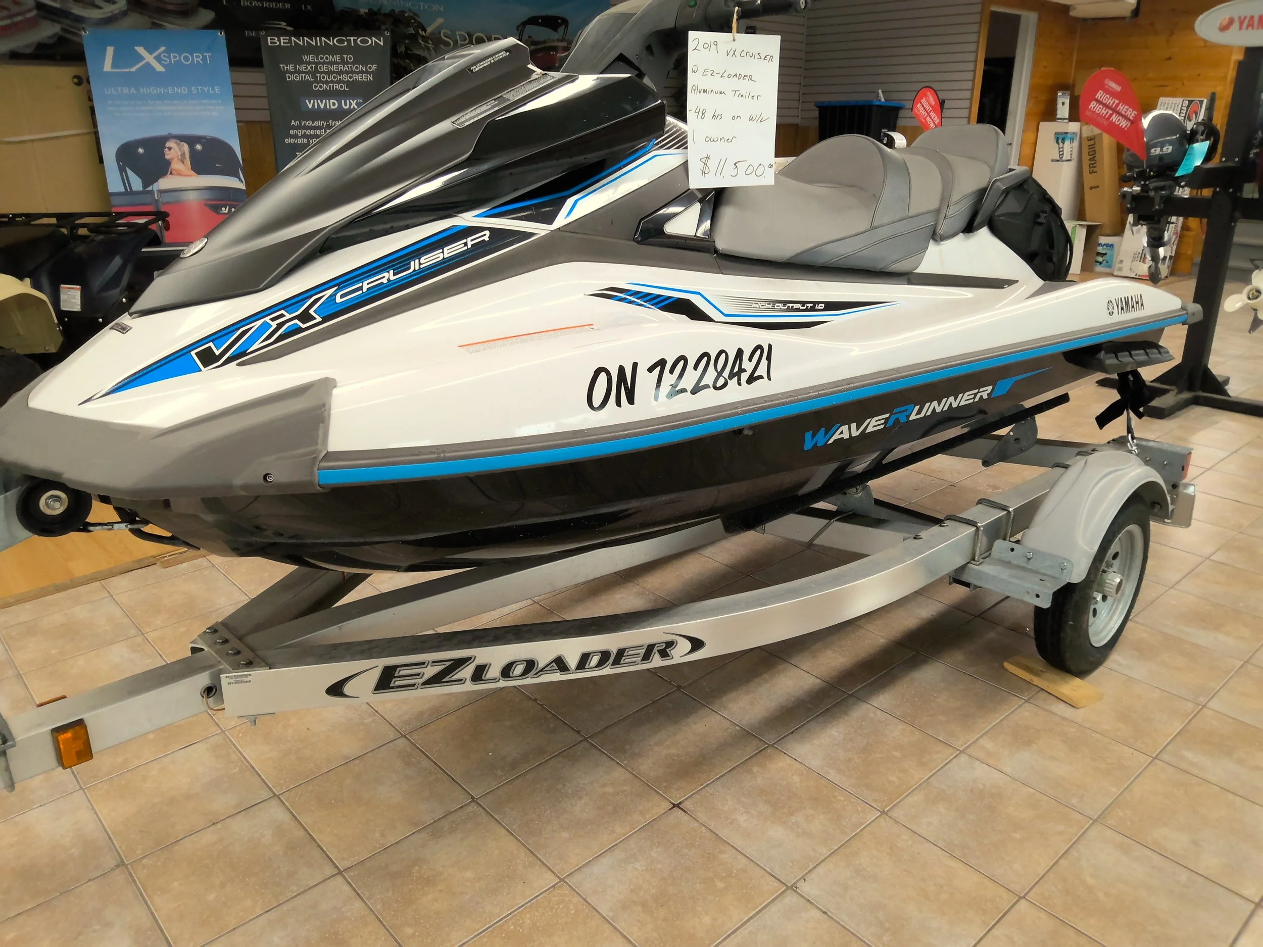A Yamaha WaveRunner jet ski on a trailer inside a store. The jet ski is mainly white with black and blue accents. There are signs in the background, including a white handwritten sign providing  for sale, which is a 2019 Yamaha VX Cruiser with an E2-Loader aluminum trailer, 48 hours on the engine, and priced at $11,500.