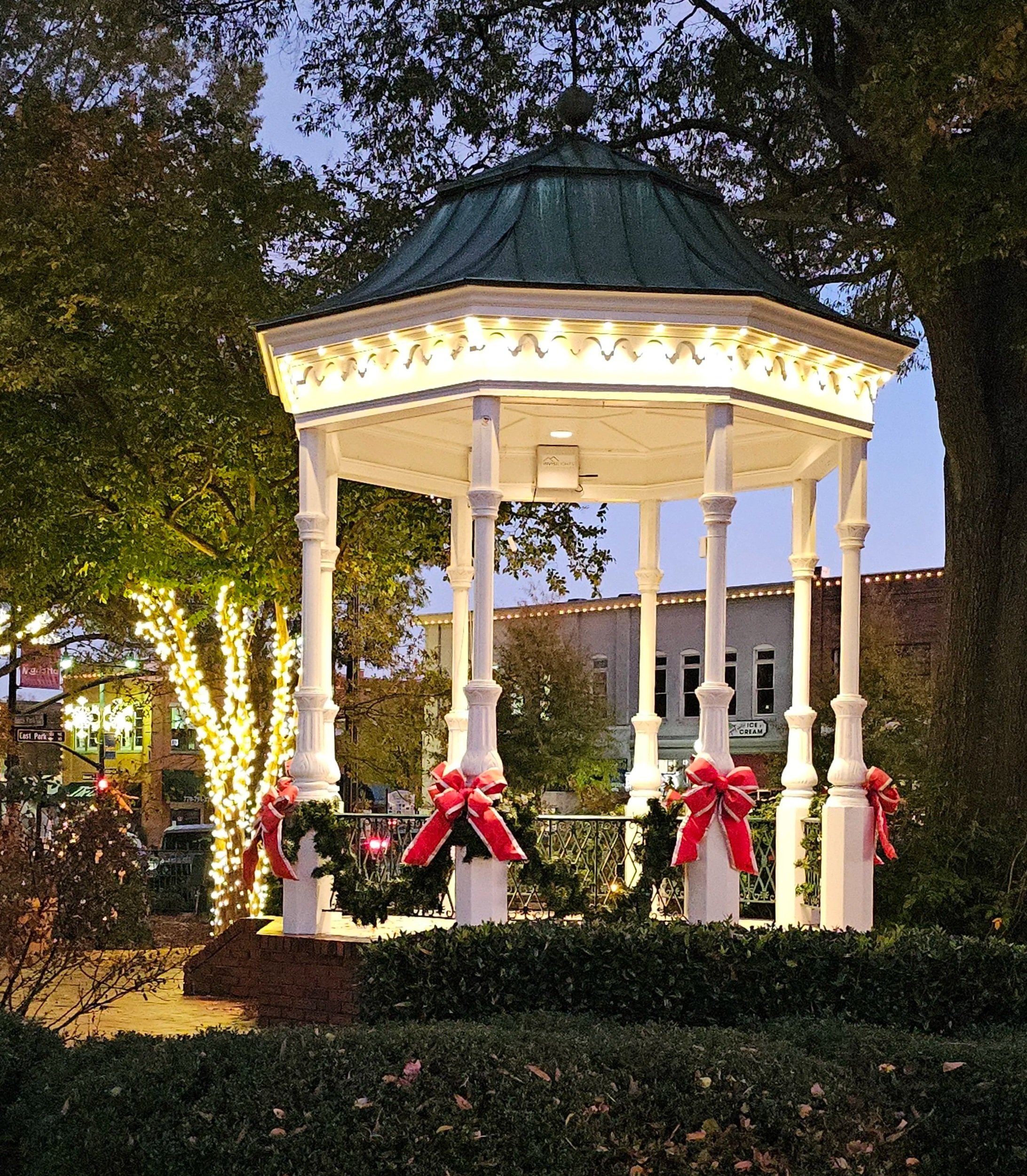 A white gazebo with decorative molding and trim, illuminated with string lights, decorated with red bows, standing in front of trees and buildings during evening time. Invisilights installed by Rich Lighting Co on Gazebo, showing classic warm white.