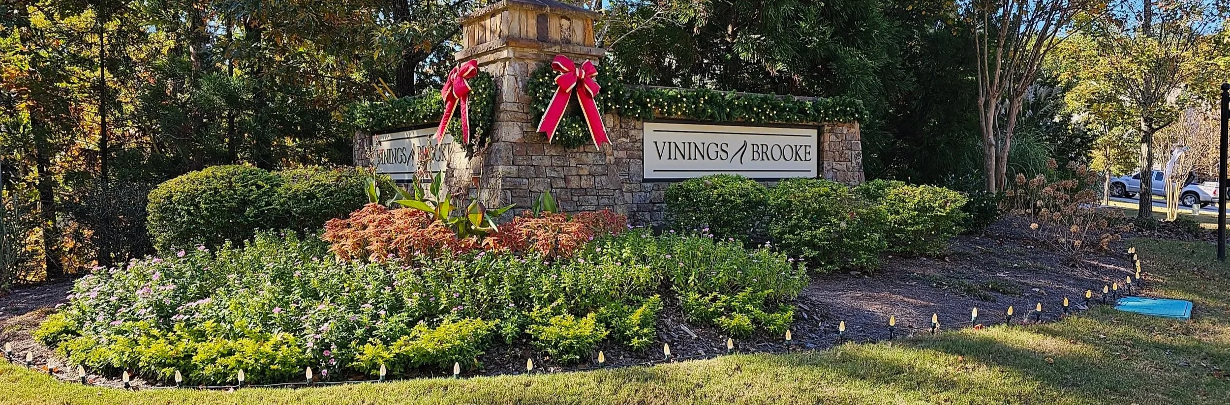 Entrance sign for Vinings Brooke surrounded by holiday decorations, greenery, and flowers, with parked cars and trees in the background.