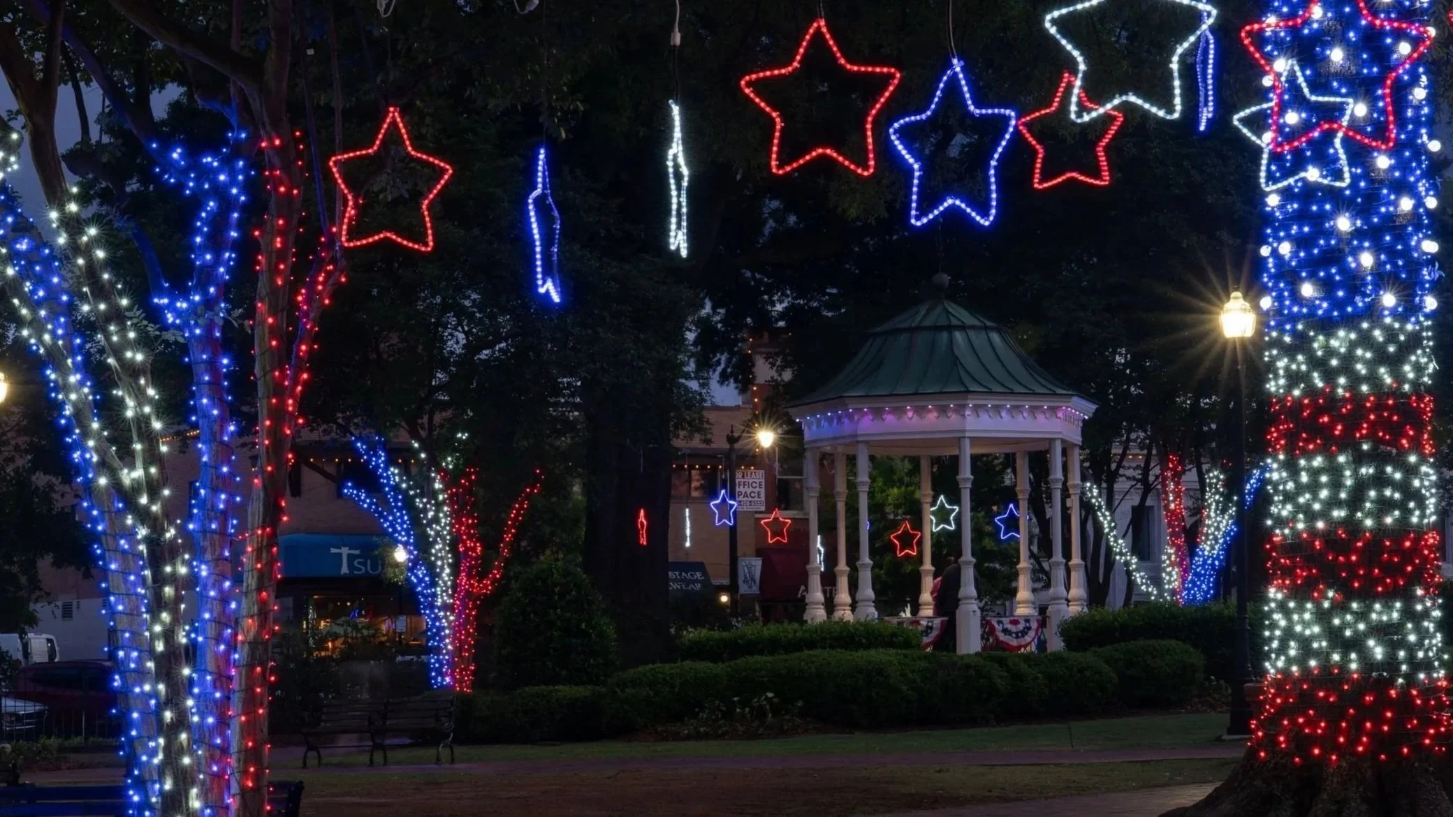 Nighttime scene of a park decorated with red, white, and blue star and stripe light displays, including illuminated star outlines hanging above, with a gazebo and streetlamp in the background.