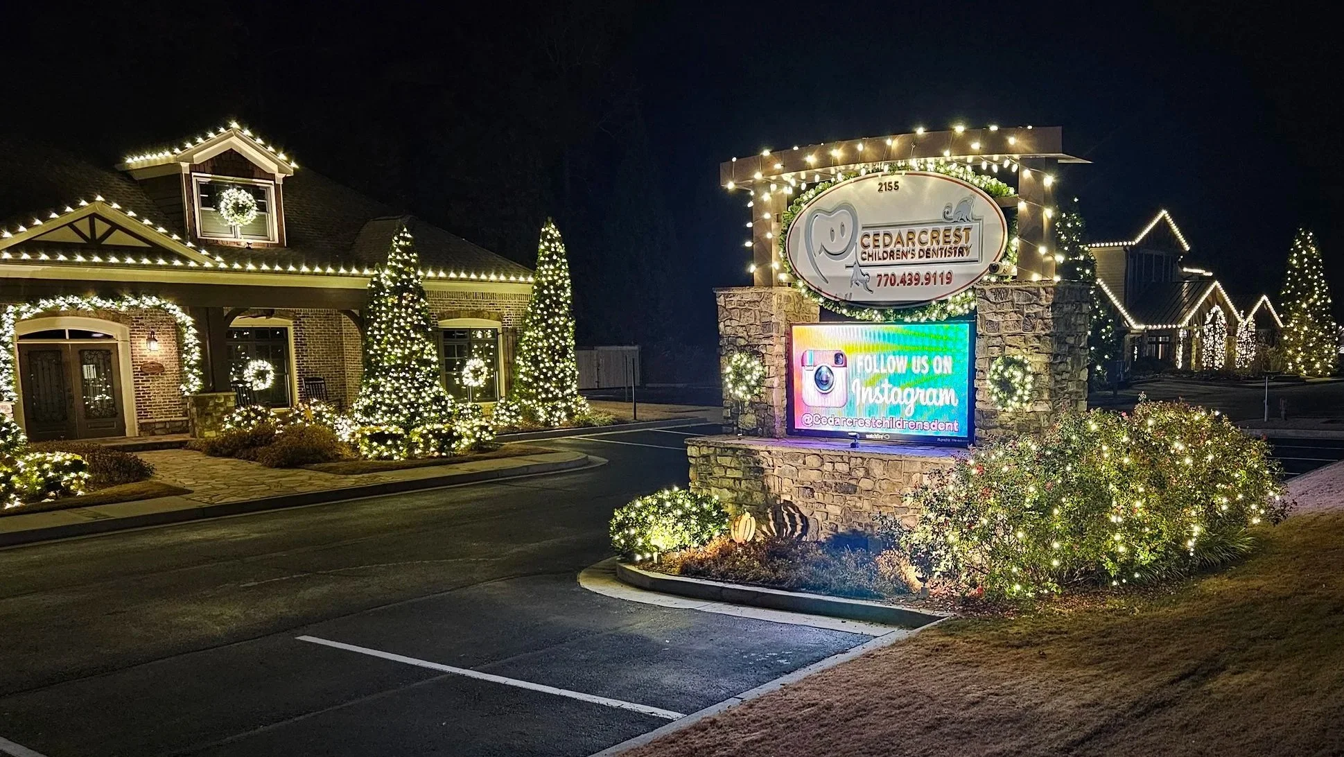 Residential building decorated with Christmas lights and wreaths. Prominent sign for Cedar Crest Children's Dentistry with social media and contact information, illuminated at night.