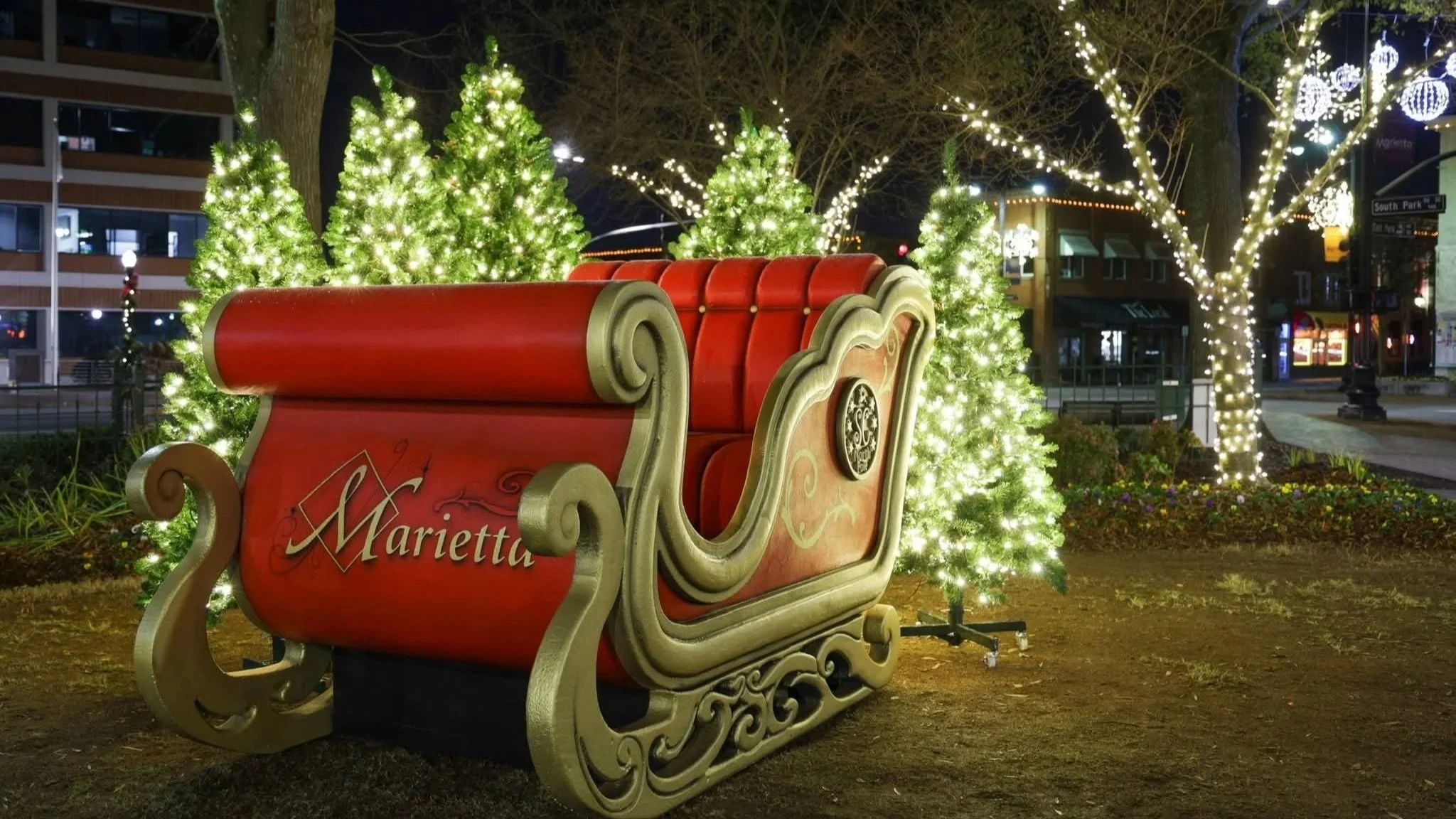 Christmas display featuring a red and gold Marietta sleigh surrounded by illuminated green Christmas trees and decorated trees with white lights in a city park at night.