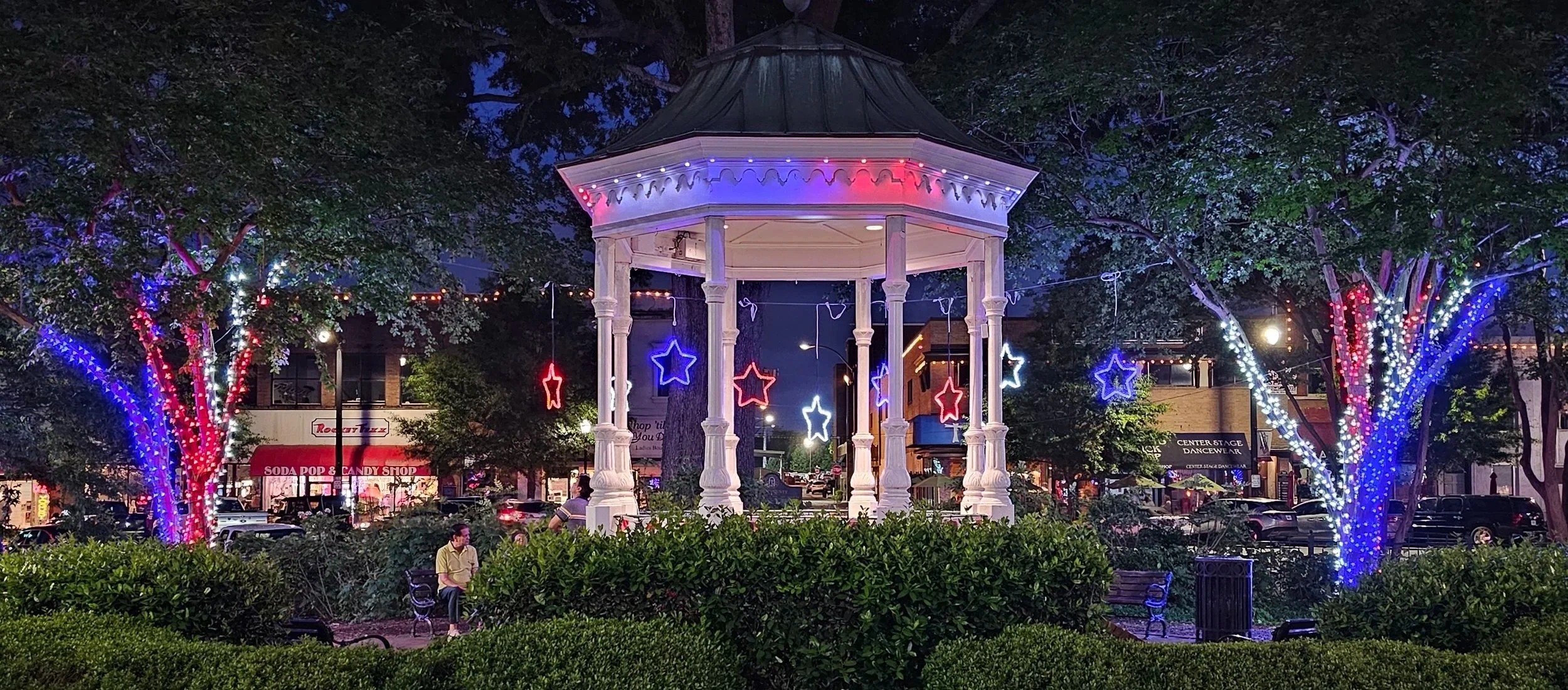 A decorative gazebo in a park illuminated with colorful string lights and star-shaped decorations at night. Surrounding trees are wrapped in blue, white, and red lights. Invisilights installed on Gazebo. Design by Rich Lighting Co for Marietta Square
