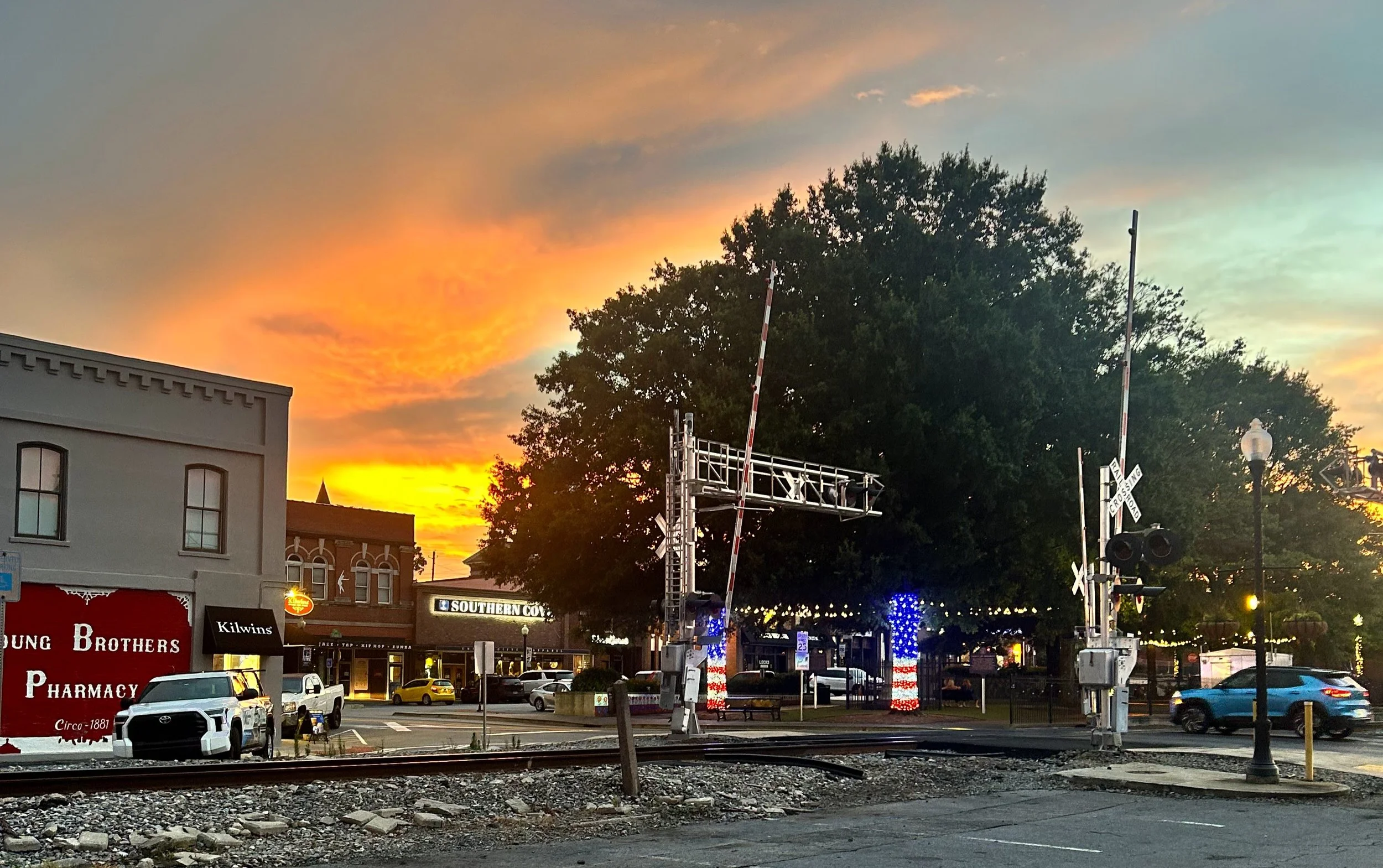 A small downtown area at sunset with a colorful sky. There are buildings, parked cars, train tracks, and a train crossing signal in the scene. The sky has shades of orange, pink, and purple with some cloud cover.