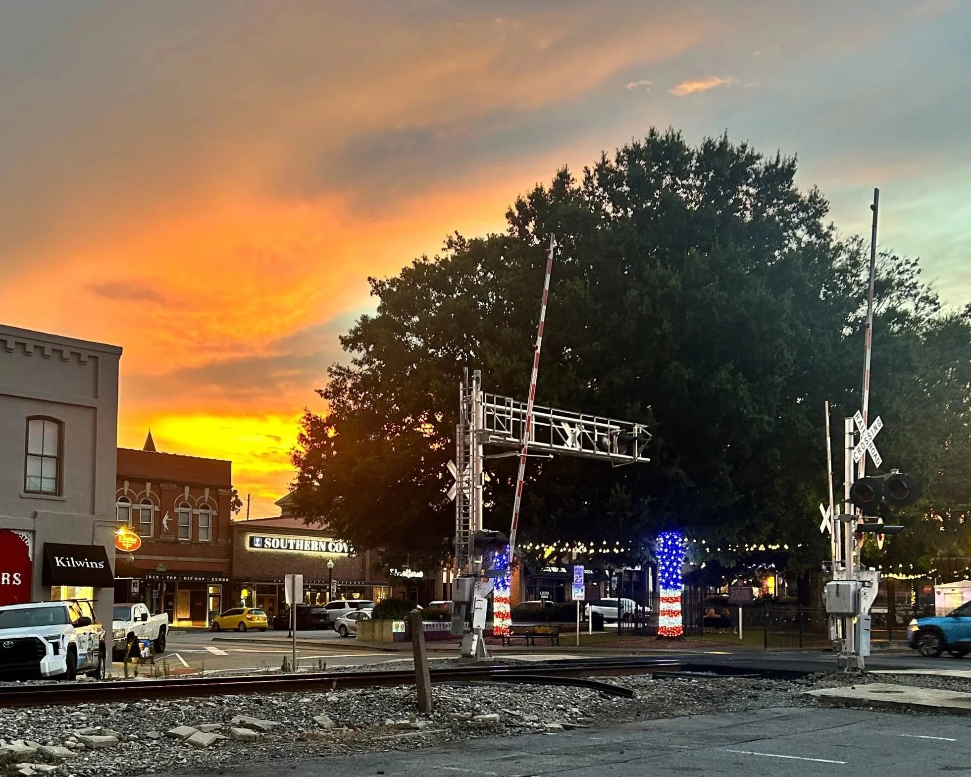 A town street scene at sunset with a colorful sky, trees, a railroad crossing, parked cars, and storefronts including a Kilwins shop and a Southern Cow restaurant.