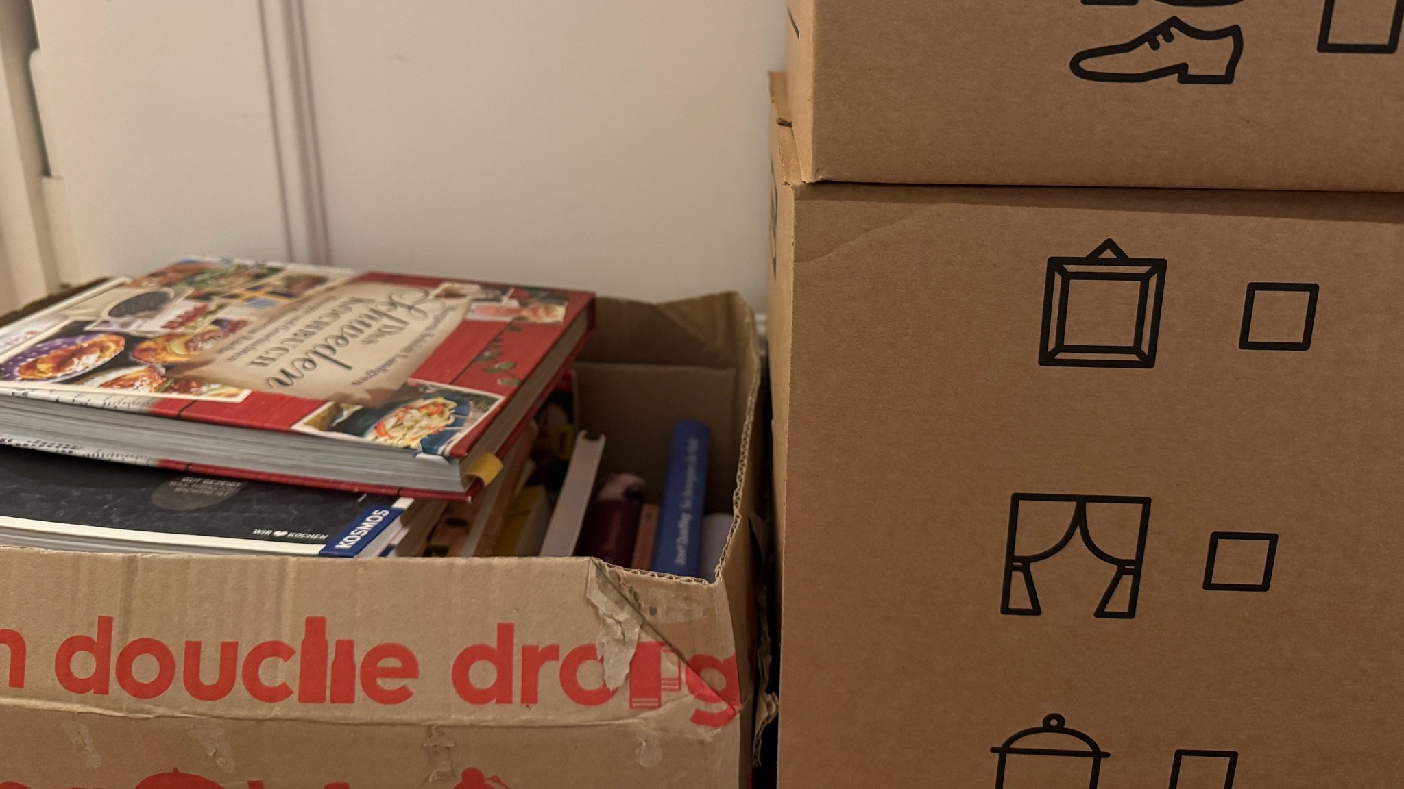 Drei Kisten, eine bis zum Rand gefüllt mit Büchern, zwei gestapelt. Ihr Inhalt ist nicht zu sehen.