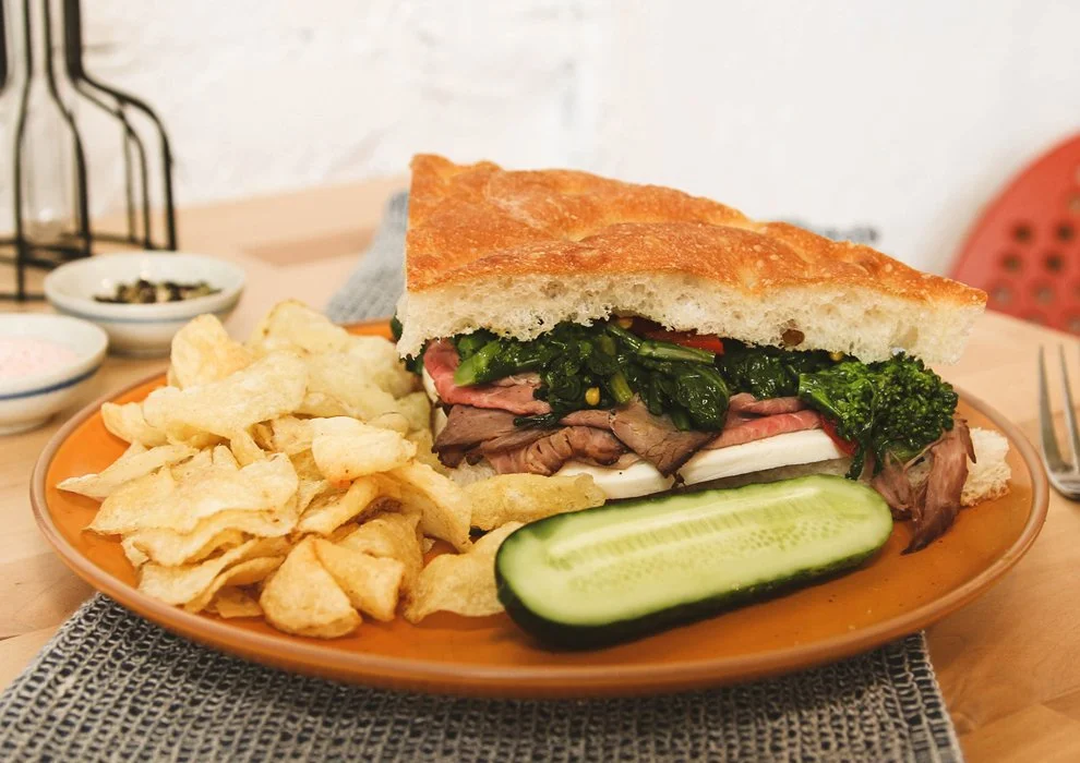 Italian Cowboy. House-made roasted beef, charred broccoli rabe, mozzarella, sweet chili peppers, roasted garlic, calabrian chili oil, on focaccia, on a plate with potato chips and a pickle.