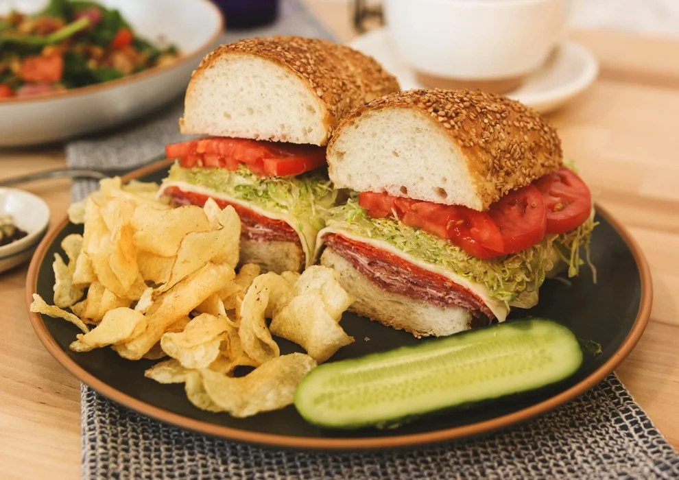 The Big Italian. Coppa, pepperoni, soppressata, provolone cheese, tomato, shredded lettuce, spicy red wine vinaigrette, on sesame seed hero, on a plate with potato chips and a pickle.