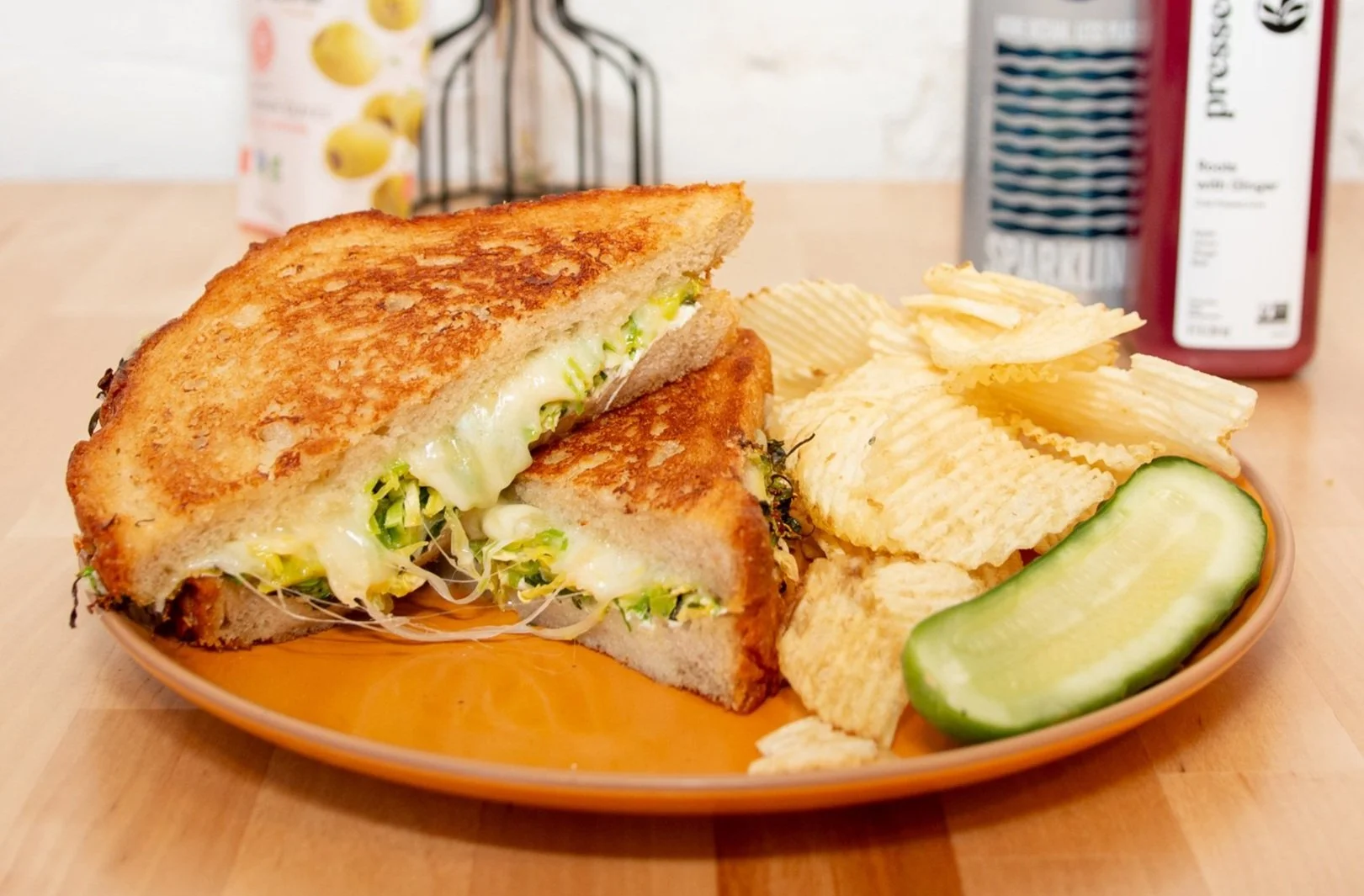 Zoller's Brussel Grilled Cheese Sandwich. Fresh brussels sprouts, mozzarella, basil, maldon salt, and lemon oil on toasted sourdough, on a plate with potato chips and a pickle.