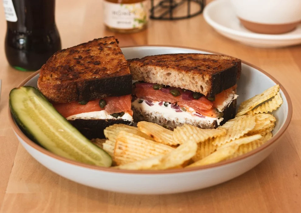 Smoked Salmon Sandwich. Salmon, whipped cream cheese, pickled pearl onion, capers, on toasted rye, on a plate with potato chips and a pickle.