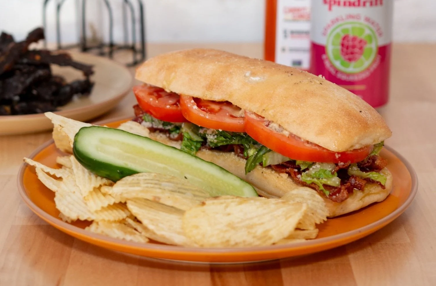 Caesar BLT. Caesar mayonnaise, bacon, romaine, tomato, on a long club roll, on a plate with potato chips and a pickle.