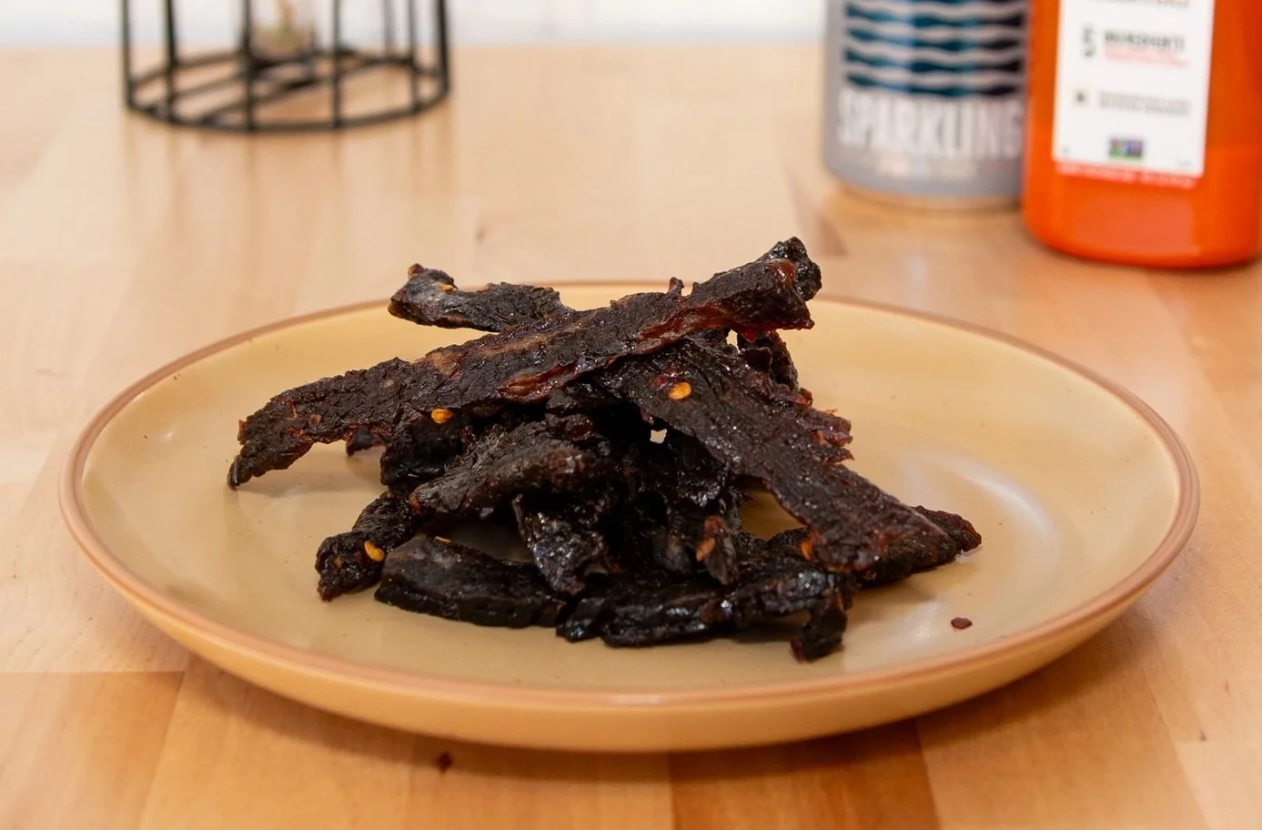 A beige plate with beef jerky strips on a wooden table, with bottles of water and juice in the background.