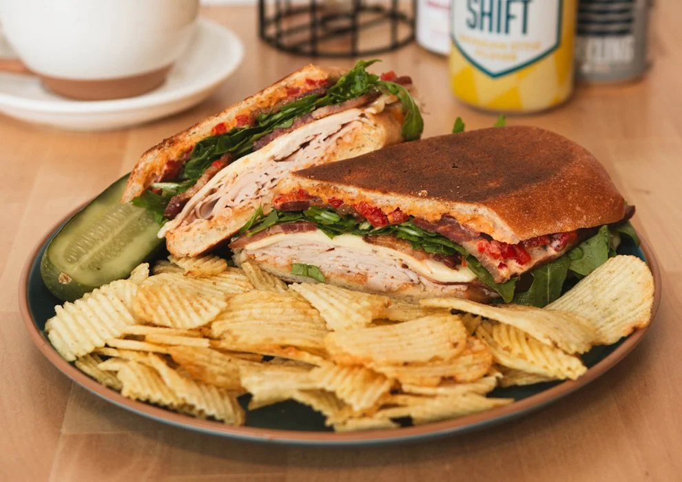 The Terrace Turker. Smoked turkey, bacon, roasted red pepper, American white cheese, arugula, terrace sauce, on a long club roll, on a plate with potato chips and a pickle.