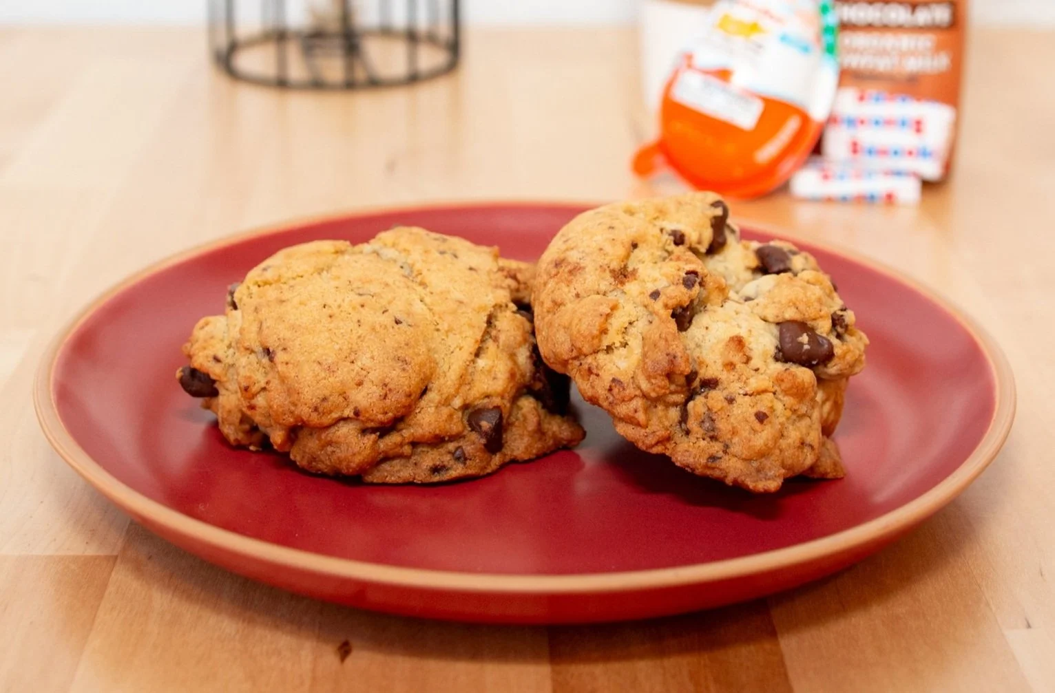 Two brown butter chocolate chip cookies on a red plate placed on a wooden table with packaged snack foods in the background.