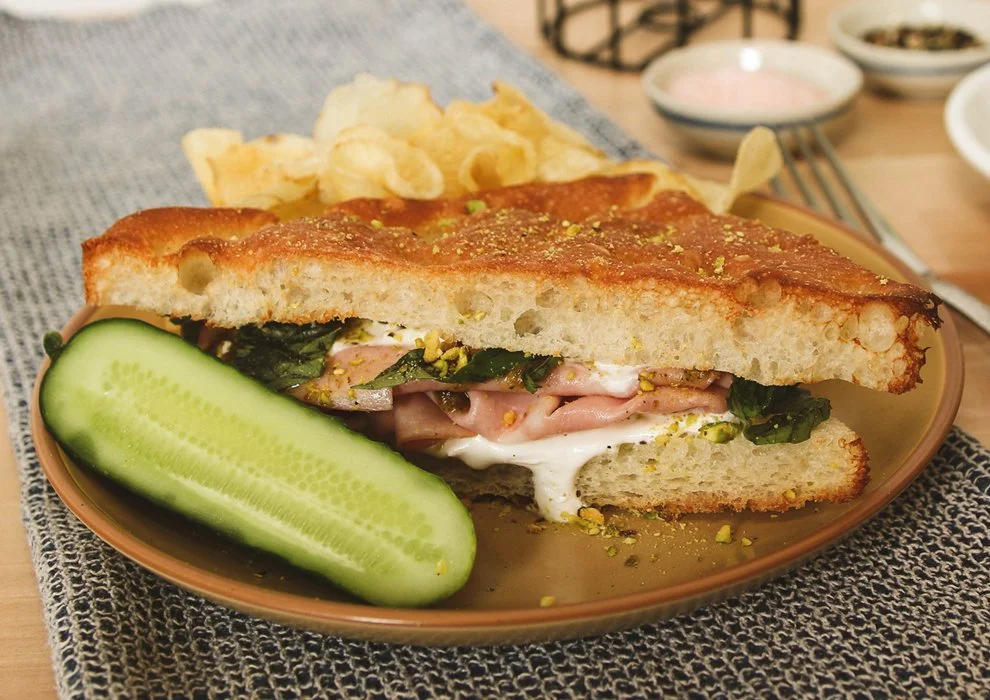 The Mort. Mortadella, stracciatella, pistachio pesto, basil, on a plate with potato chips and a pickle.