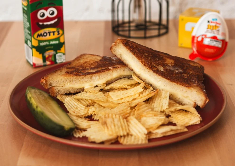 Classic Grilled Cheese. American cheese, mayonnaise, on white bread, on a plate with potato chips and a pickle.