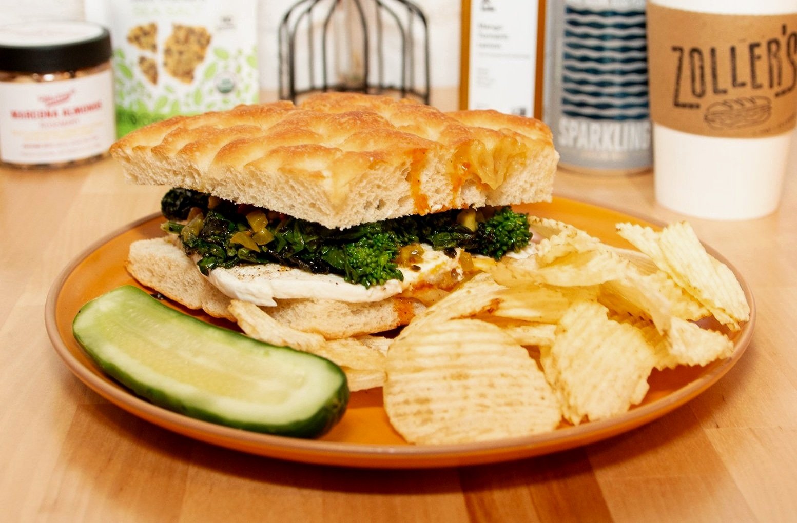 Ricci Cowboy. Charred broccoli rabe, mozzarella, sweet chili peppers, roasted garlic, calabrian chili oil, on focaccia, on a plate with potato chips and a pickle.