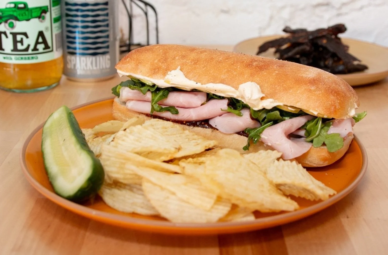 Pig & Fig. Black forest ham, brie, sliced apple, fig spread, and arugula on long club roll, on a plate with potato chips and a pickle.
